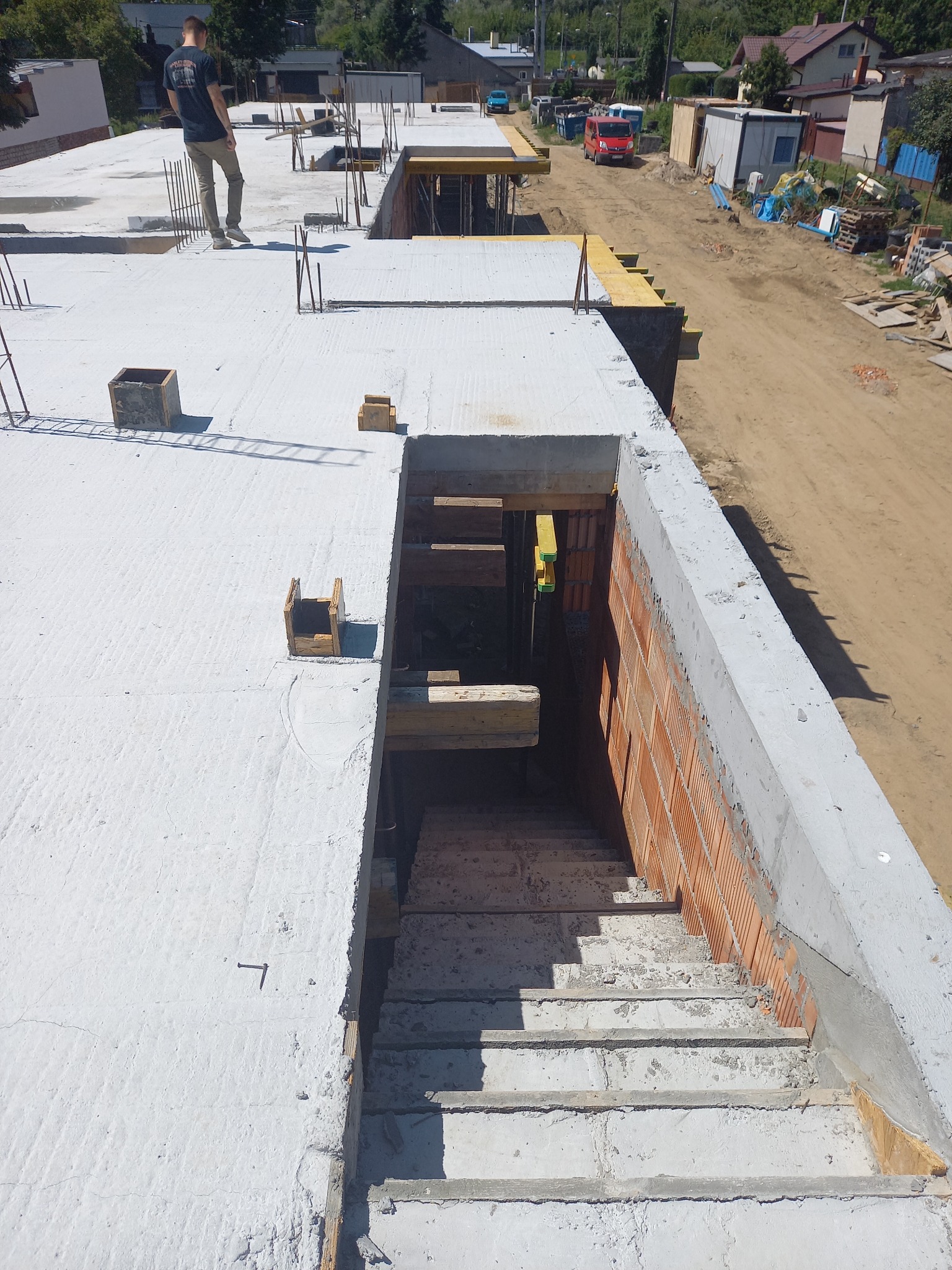 Wysoki kąt widzenia na strop żelbetowy w trakcie budowy domu jednorodzinnego, widoczne zbrojenia, szalunki, oraz fragment schodów z cegły i betonu. W tle częściowo widoczna zabudowa jednorodzinna...