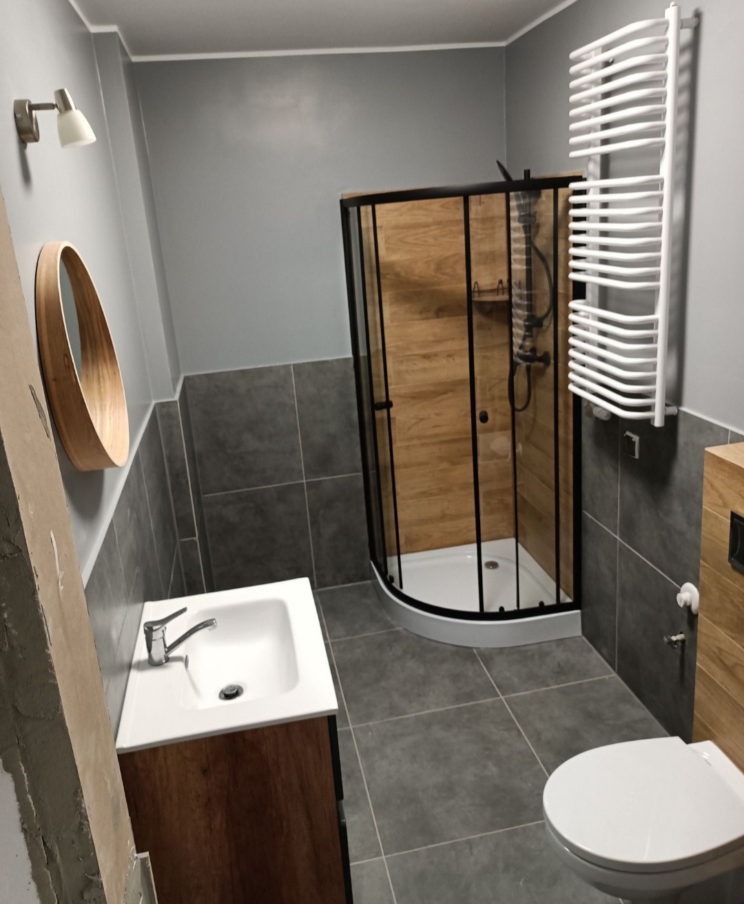 Wykończona łazienka z narożną kabiną prysznicową z czarnymi profilami i panelami imitującymi drewno, umywalką z szafką w odcieniach brązu, grzejnikiem drabinkowym oraz okrągłym lustrem z drewnianą...