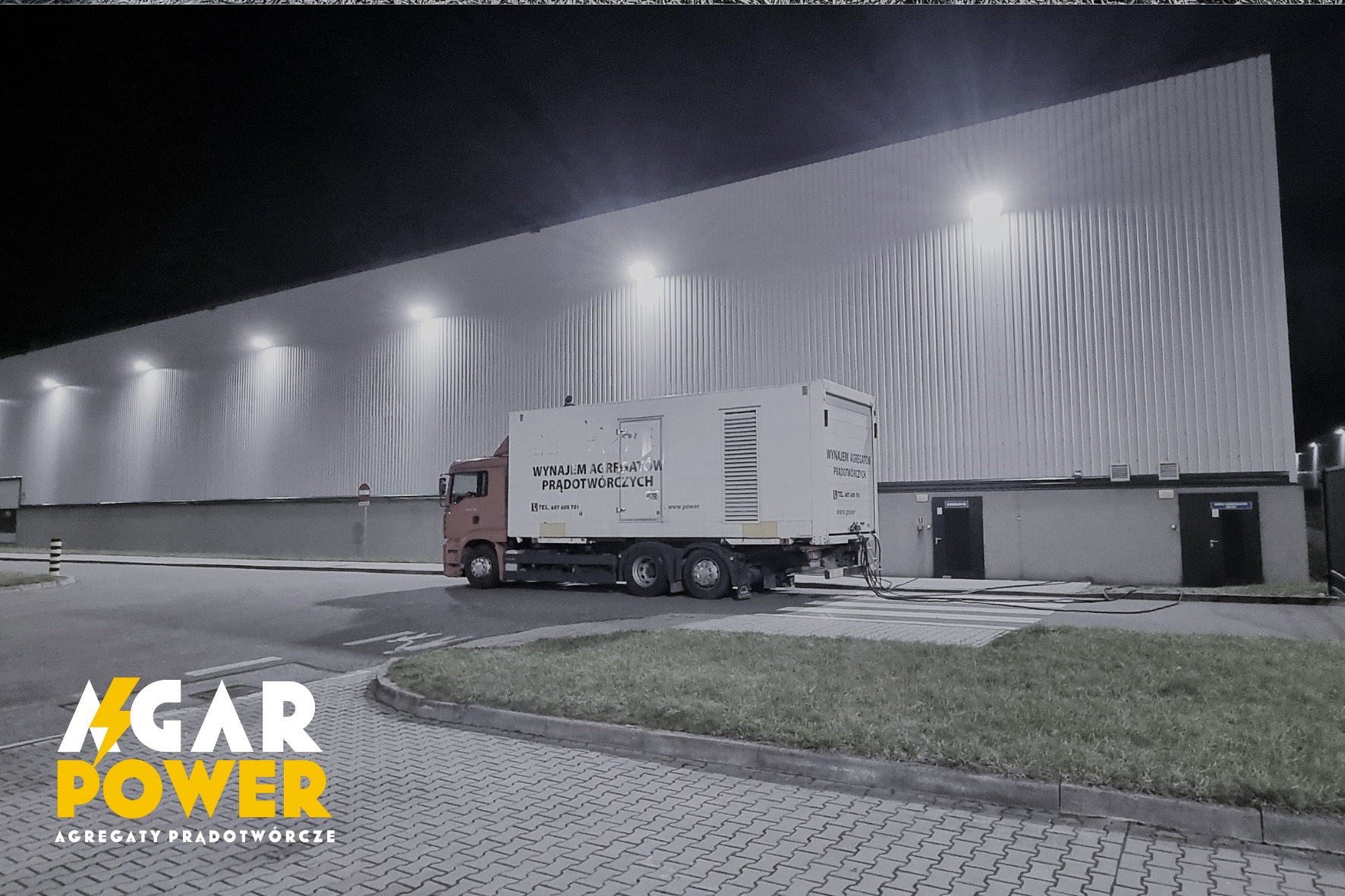 Ciężarówka z agregatem prądotwórczym na przyczepie, zaparkowana nocą przed halą przemysłową z oświetleniem zewnętrznym.