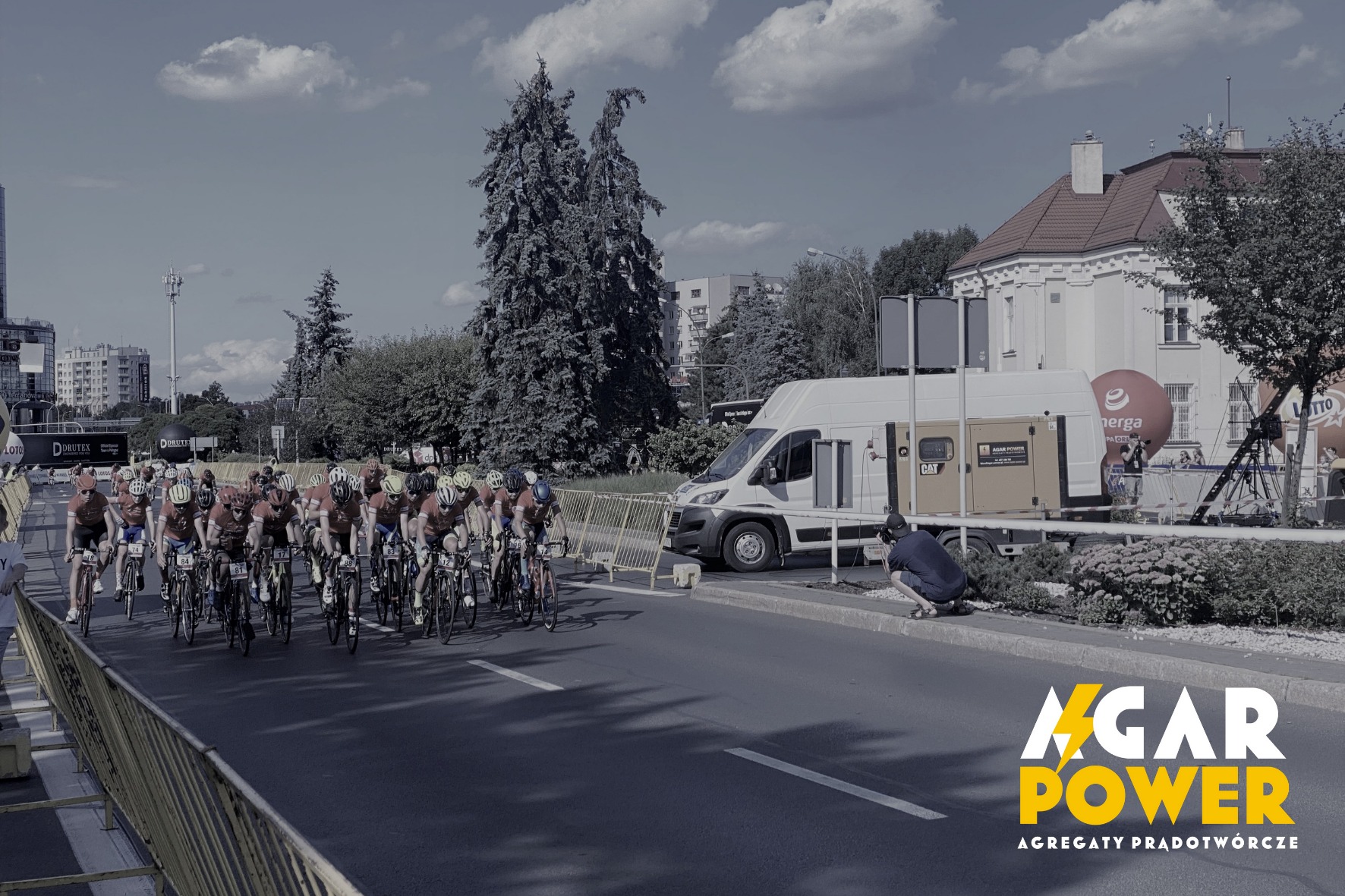 Kolarze w czerwonych koszulkach jadący ulicą, w tle biały van z agregatem prądotwórczym marki CAT, fotograf robiący zdjęcia z boku.