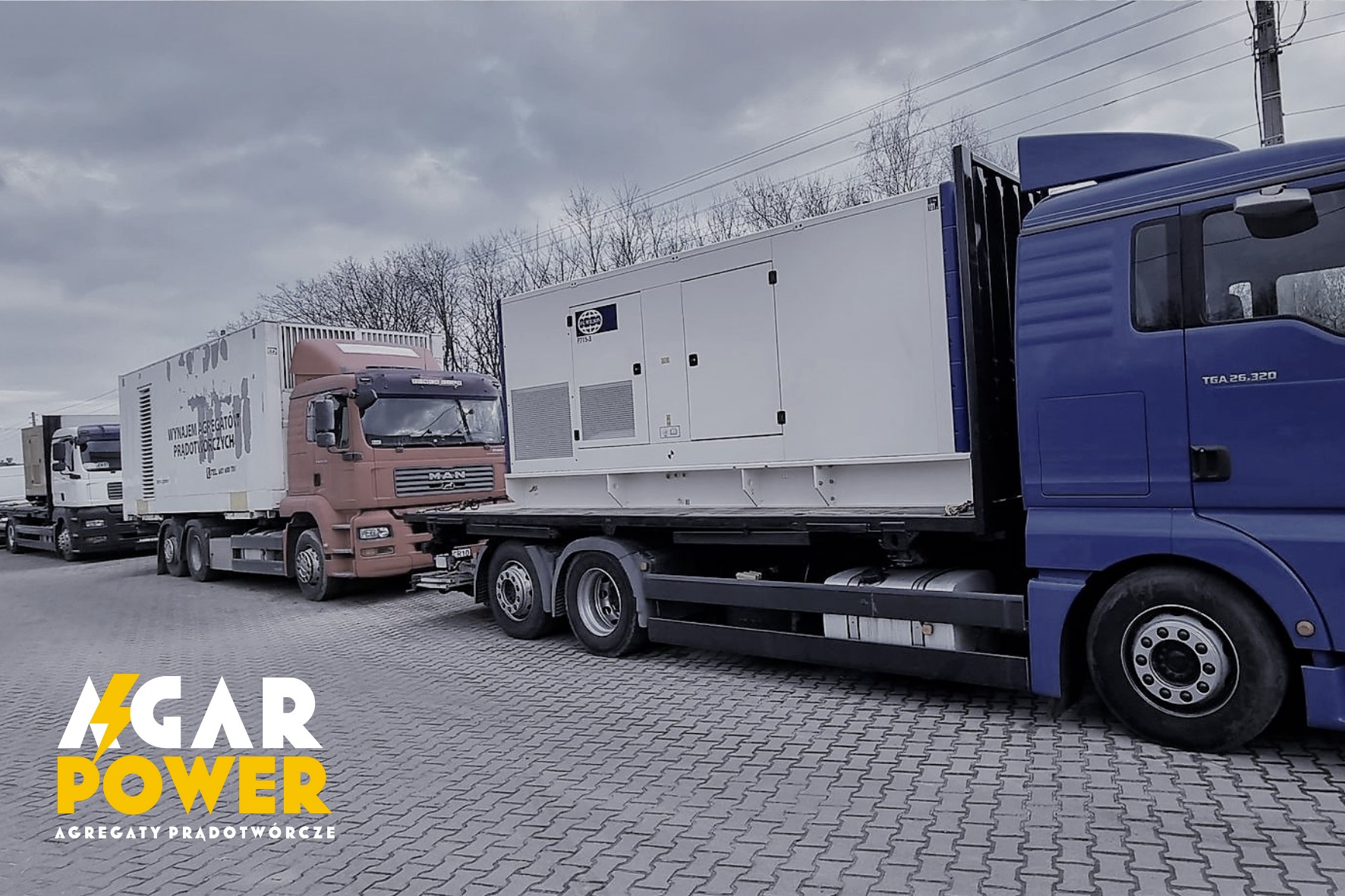 Rząd ciężarówek MAN z agregatami prądotwórczymi, widoczny logo firmy na pierwszym planie, szara kostka brukowa i pochmurne niebo.