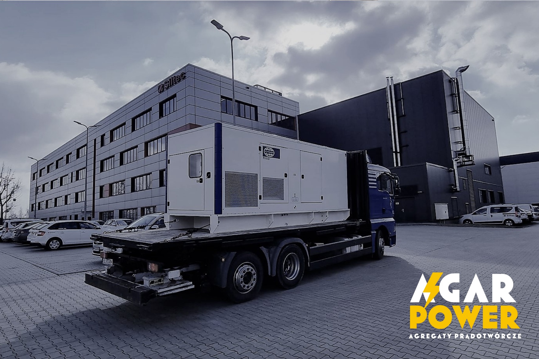 Agregat prądotwórczy na lawecie transportowej w Łodzi, widoczny na tle budynku biurowego z zaparkowanymi samochodami.