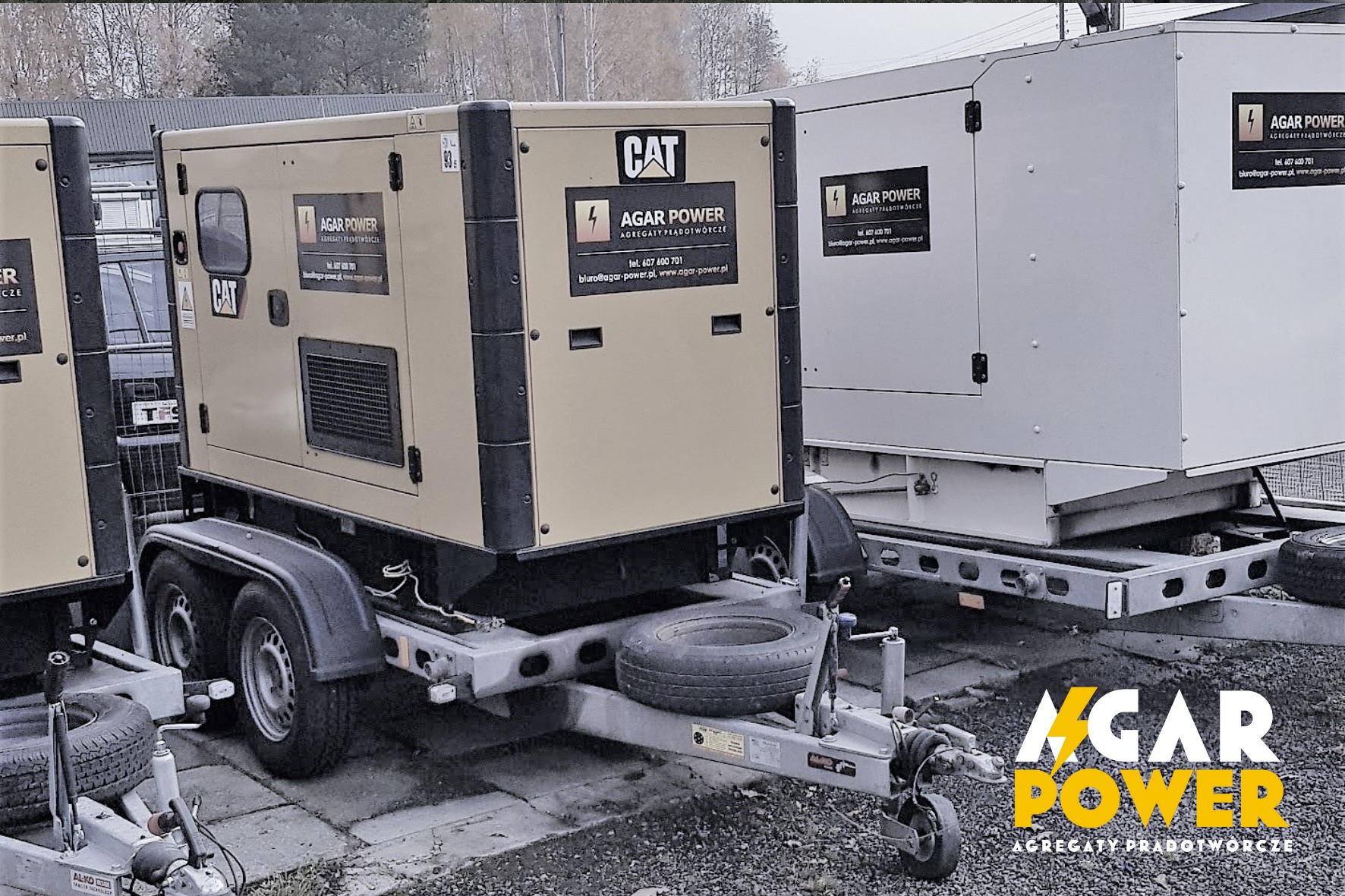 Dwa agregaty prądotwórcze na przyczepach, jeden beżowy z logo CAT, drugi biały, oba z logo Agar Power, stojące na betonowym placu.