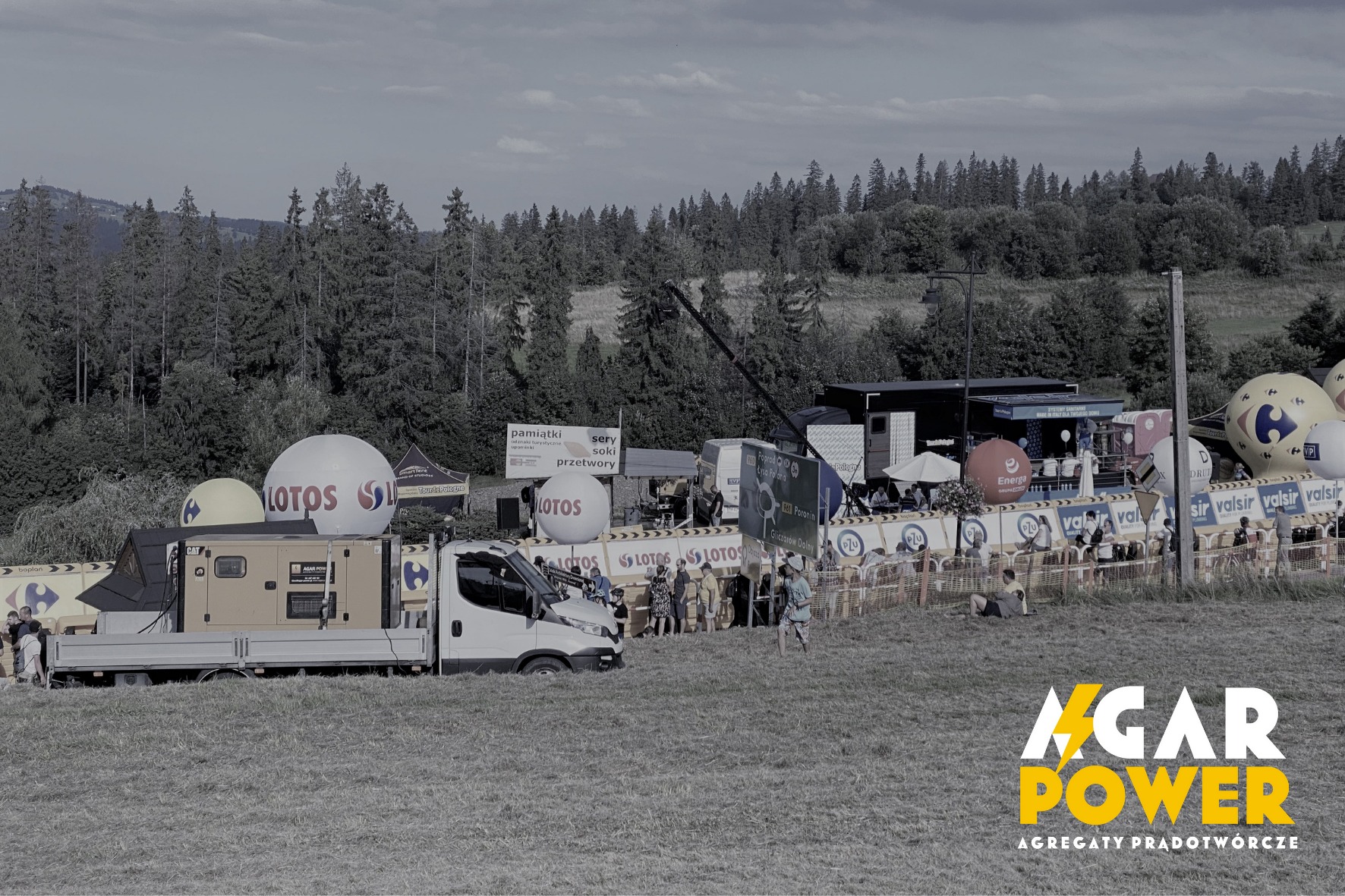 Generator prądu marki CAT na platformie samochodu dostawczego podczas imprezy plenerowej, w tle stoiska promocyjne z balonami i barierkami.