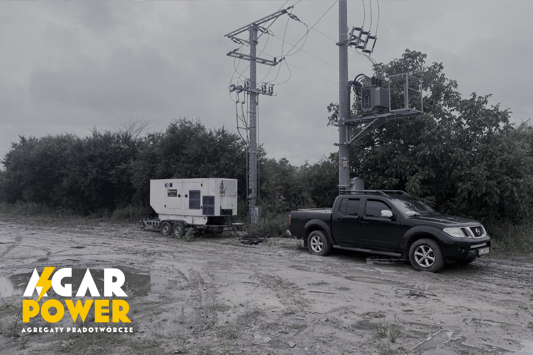 Czarny pickup Nissan Navara stoi na błotnistym terenie obok białego agregatu prądotwórczego na przyczepie, w tle słupy wysokiego napięcia i zieleń.
