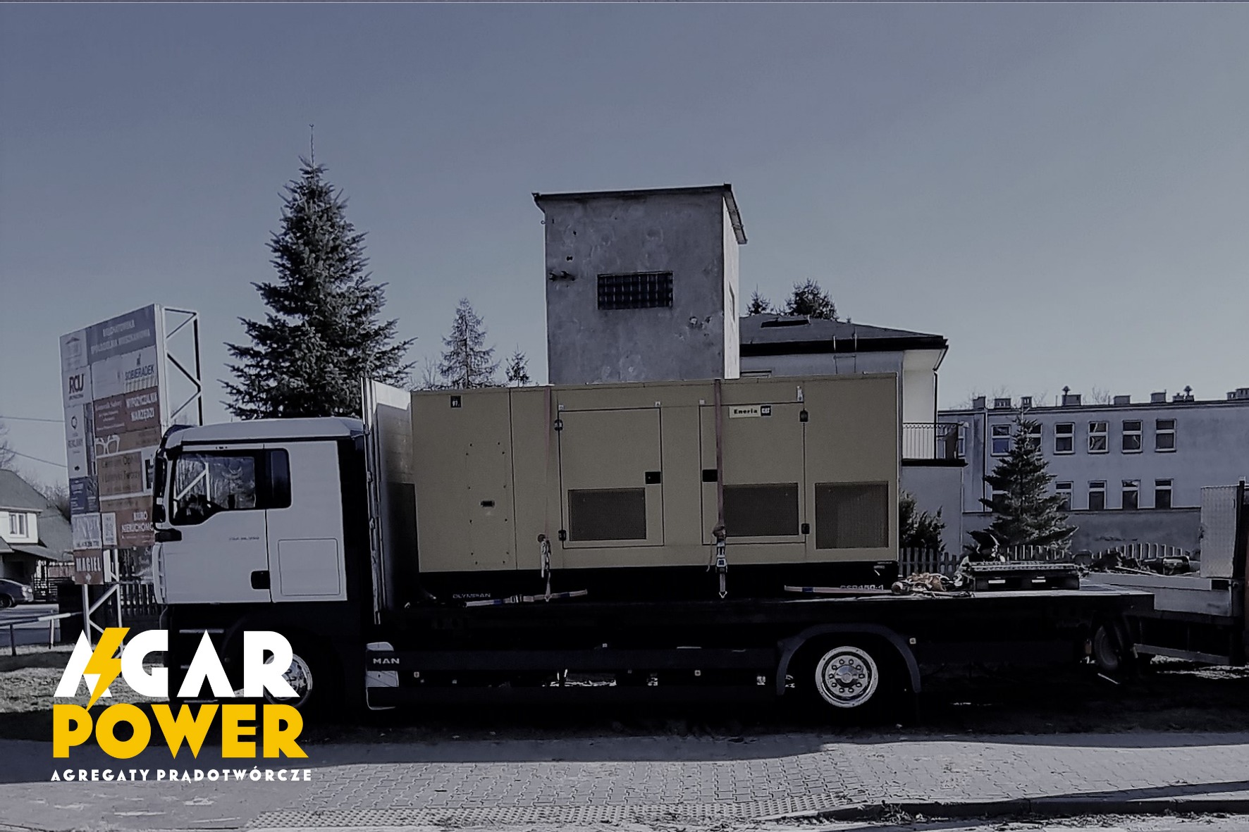 Agregat prądotwórczy marki Eneria Cat przewożony na lawecie samochodu ciężarowego MAN, na tle zabudowań miejskich i tablic reklamowych.