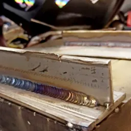 Spoina na metalowym profilu, widoczne tęczowe przebarwienia termiczne i czarna maska spawalnicza Savage w tle.