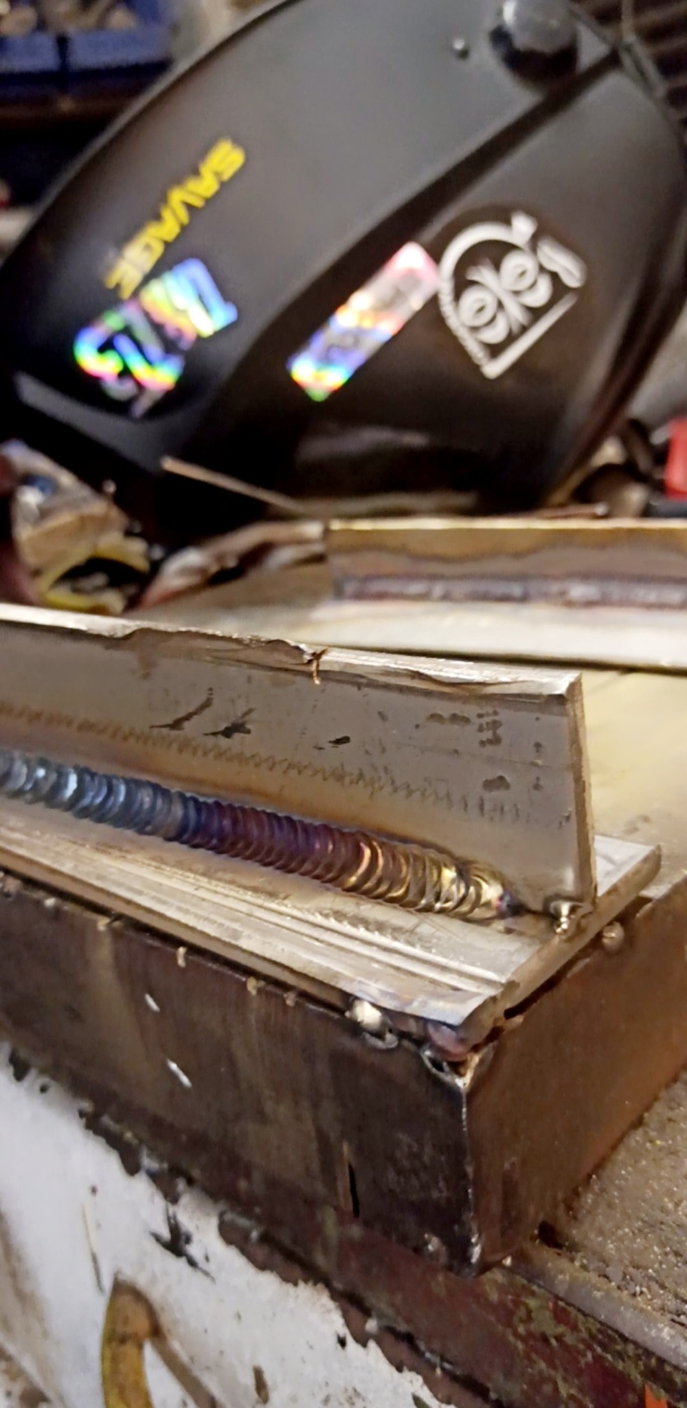 Spoina na metalowym profilu, widoczne tęczowe przebarwienia termiczne i czarna maska spawalnicza Savage w tle.