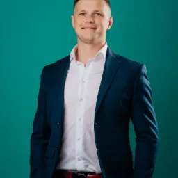 Uśmiechnięty mężczyzna w granatowym garniturze i białej koszuli, z rękami w kieszeniach, na jednolitym turkusowym tle.