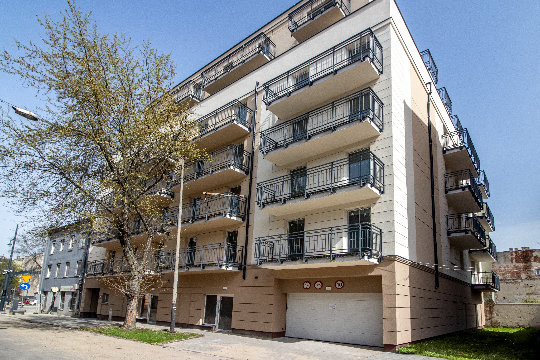 Nowoczesny, wielopiętrowy budynek mieszkalny z balkonami i garażem, widziany z poziomu ulicy w słoneczny dzień. Obok budynku rośnie drzewo.