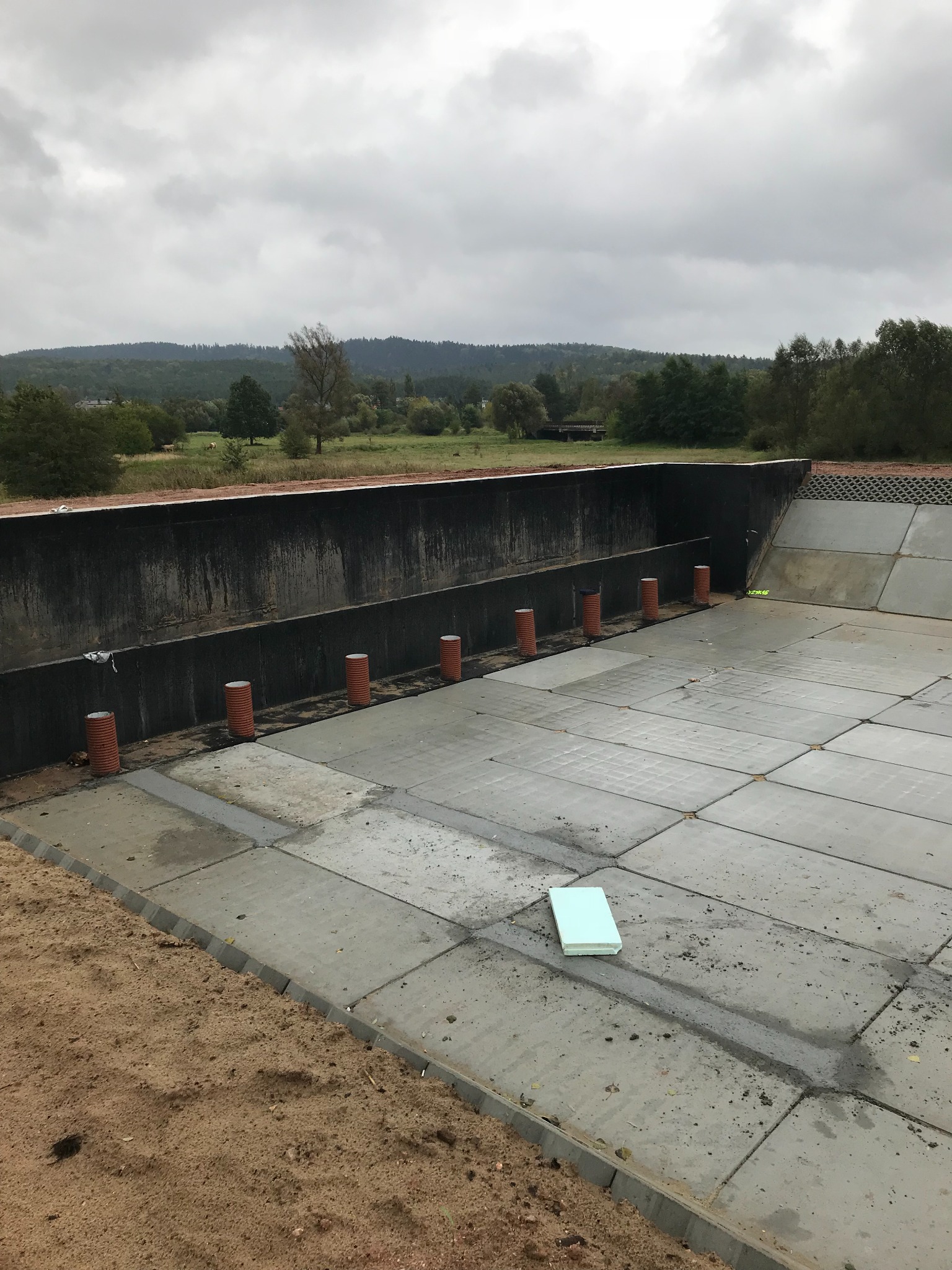 Betonowa konstrukcja w trakcie budowy z wystającymi rurami i elementem izolacyjnym, na tle krajobrazu zalesionych wzgórz pod pochmurnym niebem.