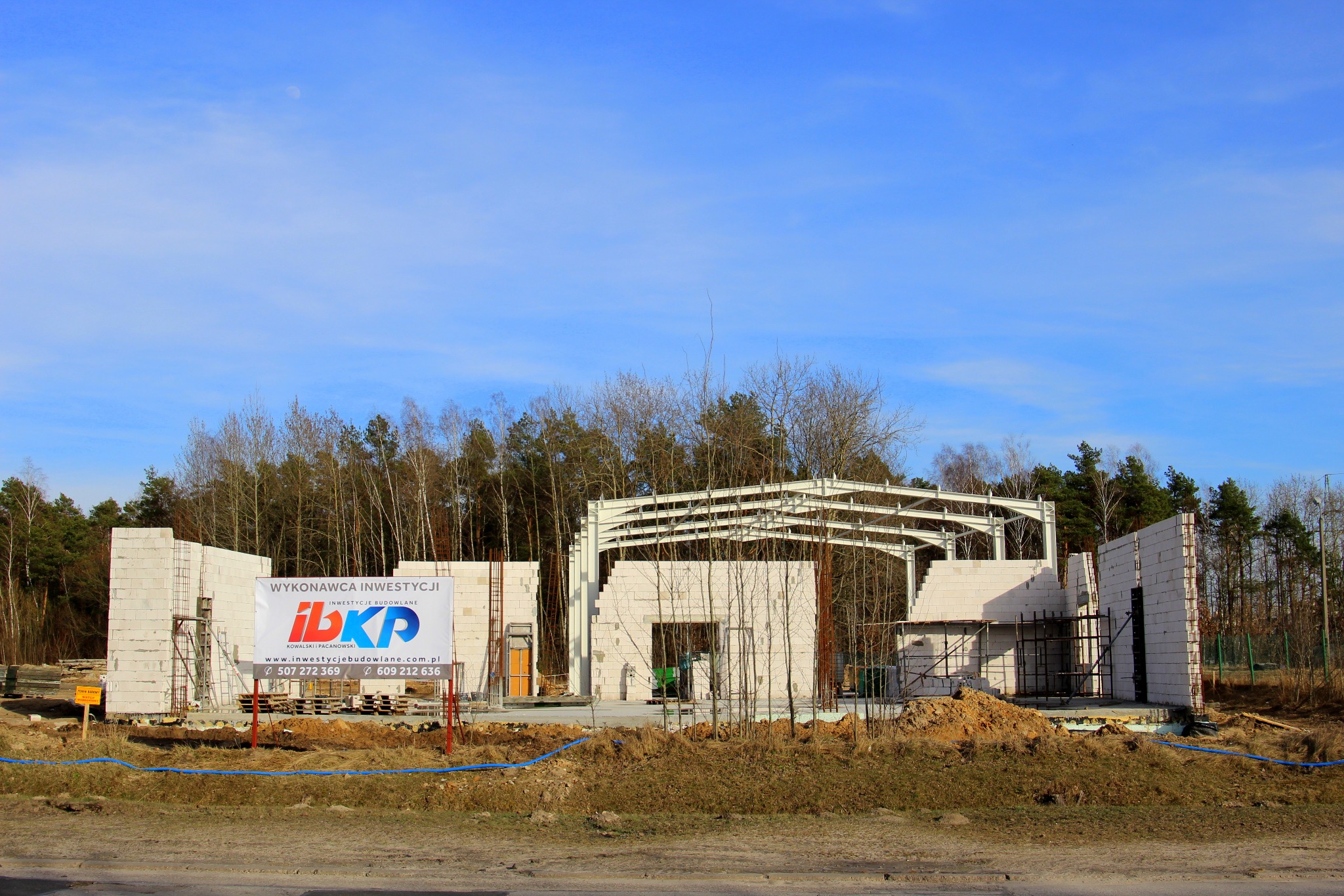 Budowa hali w toku: wzniesione ściany z betonu komórkowego, stalowa konstrukcja dachu, tablica informacyjna z logo firmy budowlanej na pierwszym planie, błękitne niebo.