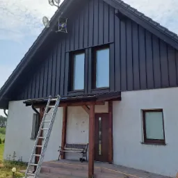Dom jednorodzinny z ciemną, pionową okładziną dachu i ścian, z zamontowaną anteną satelitarną na szczycie, drabina oparta o ścianę, widoczne ślady po pracach budowlanych.