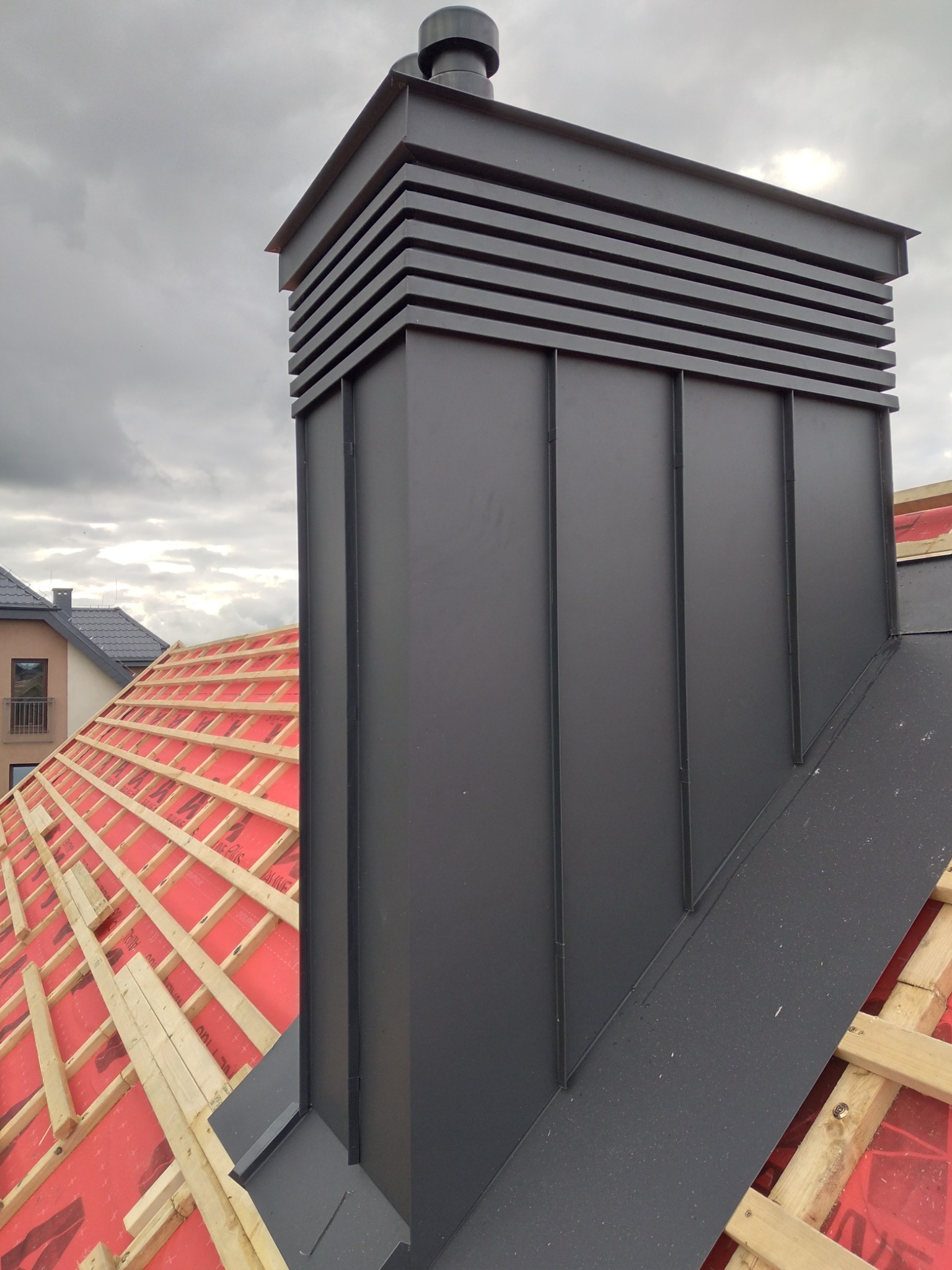 Nowy, grafitowy komin wentylacyjny z blachy na dachu w trakcie budowy, widoczne drewniane elementy konstrukcji dachu pokryte czerwoną membraną, pochmurne niebo w tle.