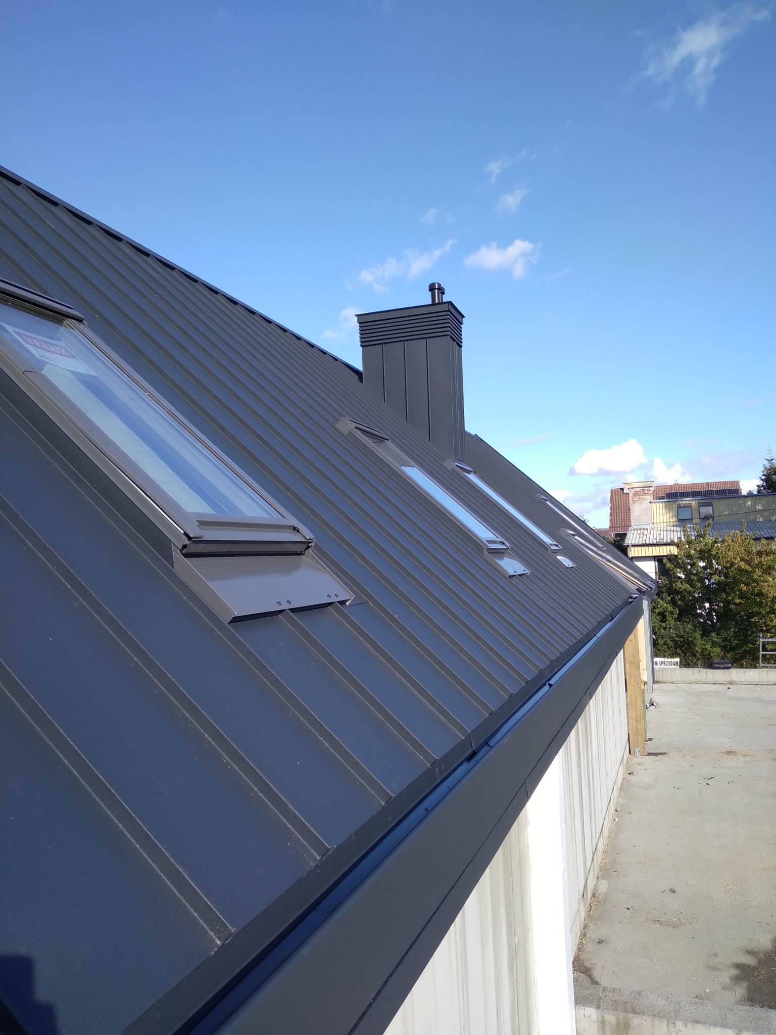 Grafitowy dach z blachy trapezowej z czterema oknami dachowymi i kominem wentylacyjnym, widok z dołu w słoneczny dzień.