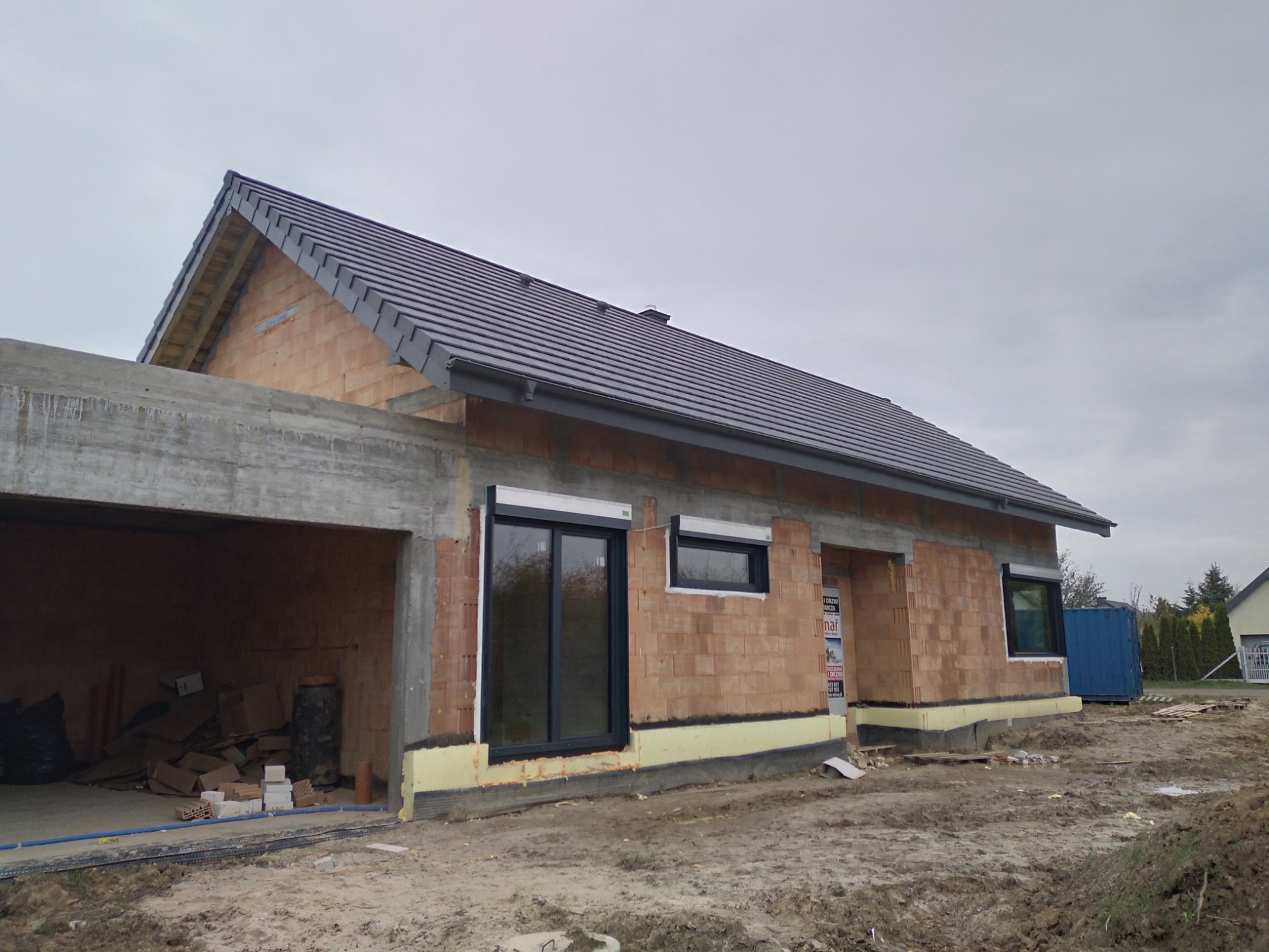 Dom w trakcie budowy z widocznym, częściowo ukończonym dachem z ciemnej dachówki i surową konstrukcją garażu; teren wokół budynku niezagospodarowany.