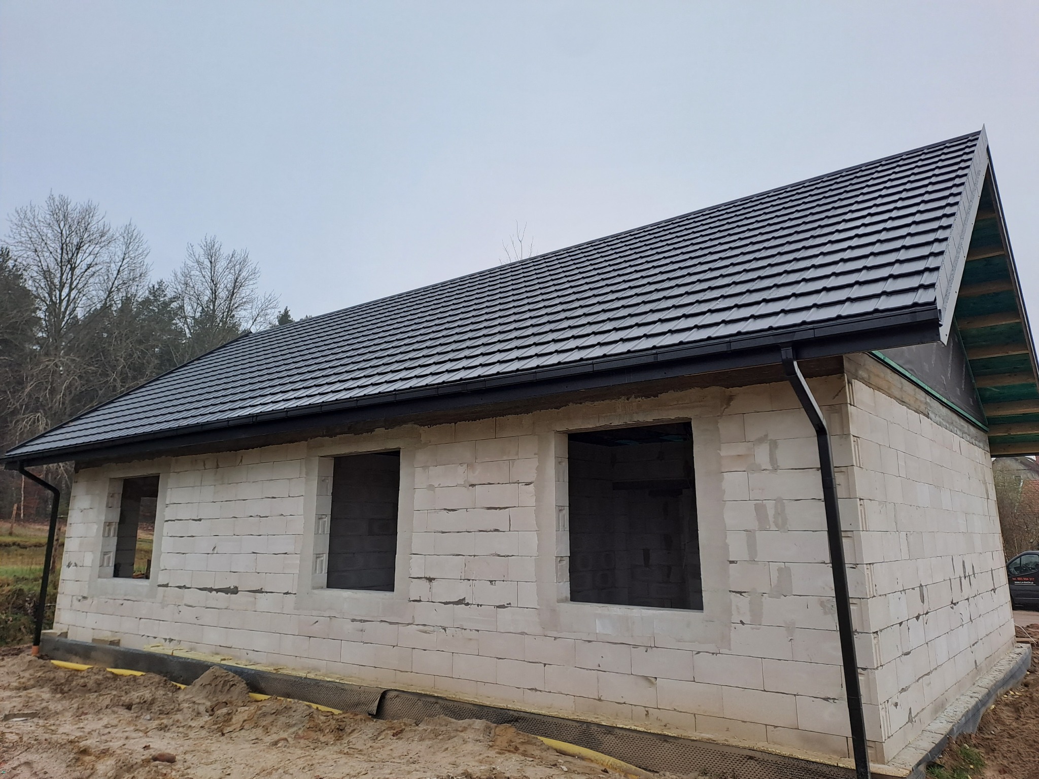 Dom w trakcie budowy z wykończonym, ciemnym dachem z dachówki modułowej i zamontowanymi rynnami, ściany z białego bloczka, widoczne otwory okienne, teren wokół domu częściowo wyrównany.