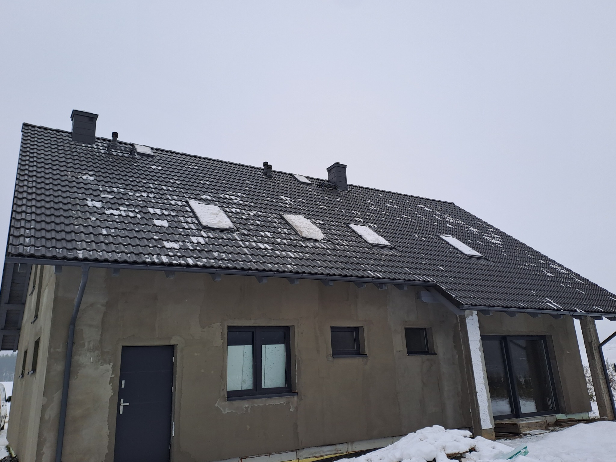 Dom w trakcie budowy z ciemną dachówką ceramiczną, widoczne okna dachowe i kominy, elewacja w stanie surowym, śnieg na dachu i wokół budynku.