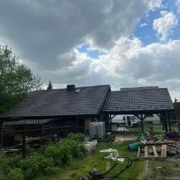 Dom jednorodzinny z nowym, ciemnym dachem z dachówki metalowej, widoczny z perspektywy ogrodu z kosiarką i elementami składowanymi na posesji, na tle pochmurnego nieba.