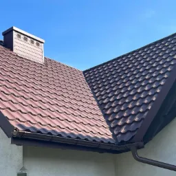 Brązowy dach z blachy trapezowej z kominem i rynnami, widok z dołu na tle błękitnego nieba.