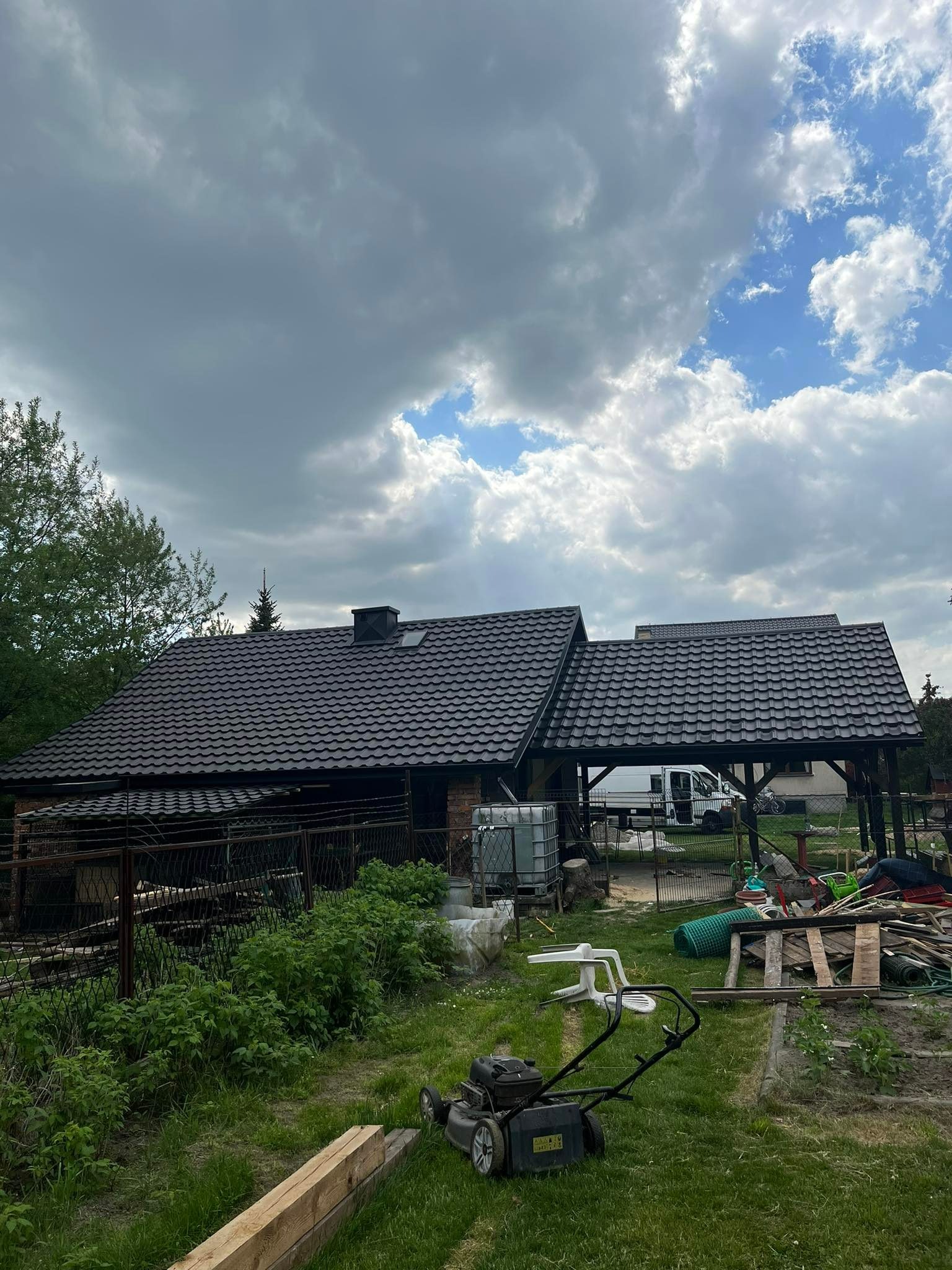 Dom jednorodzinny z nowym, ciemnym dachem z dachówki metalowej, widoczny z perspektywy ogrodu z kosiarką i elementami składowanymi na posesji, na tle pochmurnego nieba.