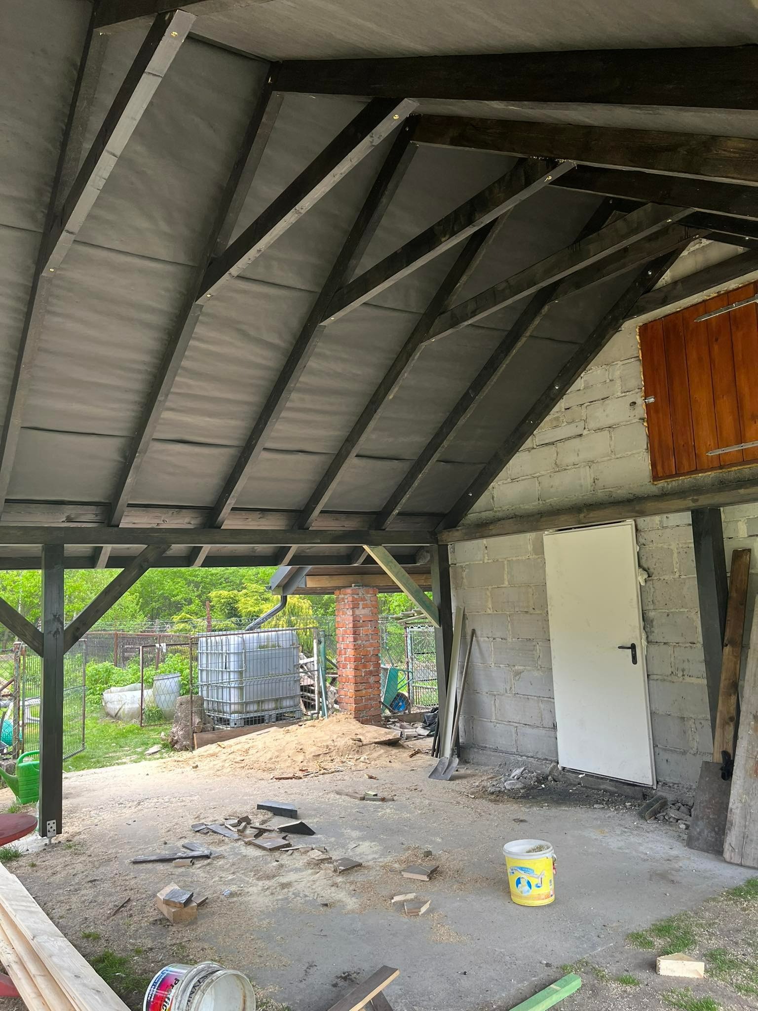 Wnętrze zadaszonej konstrukcji w trakcie budowy, widoczne drewniane elementy konstrukcyjne dachu, ściana z bloczków betonowych z białą drzwiami, oraz narzędzia i materiały budowlane na ziemi.