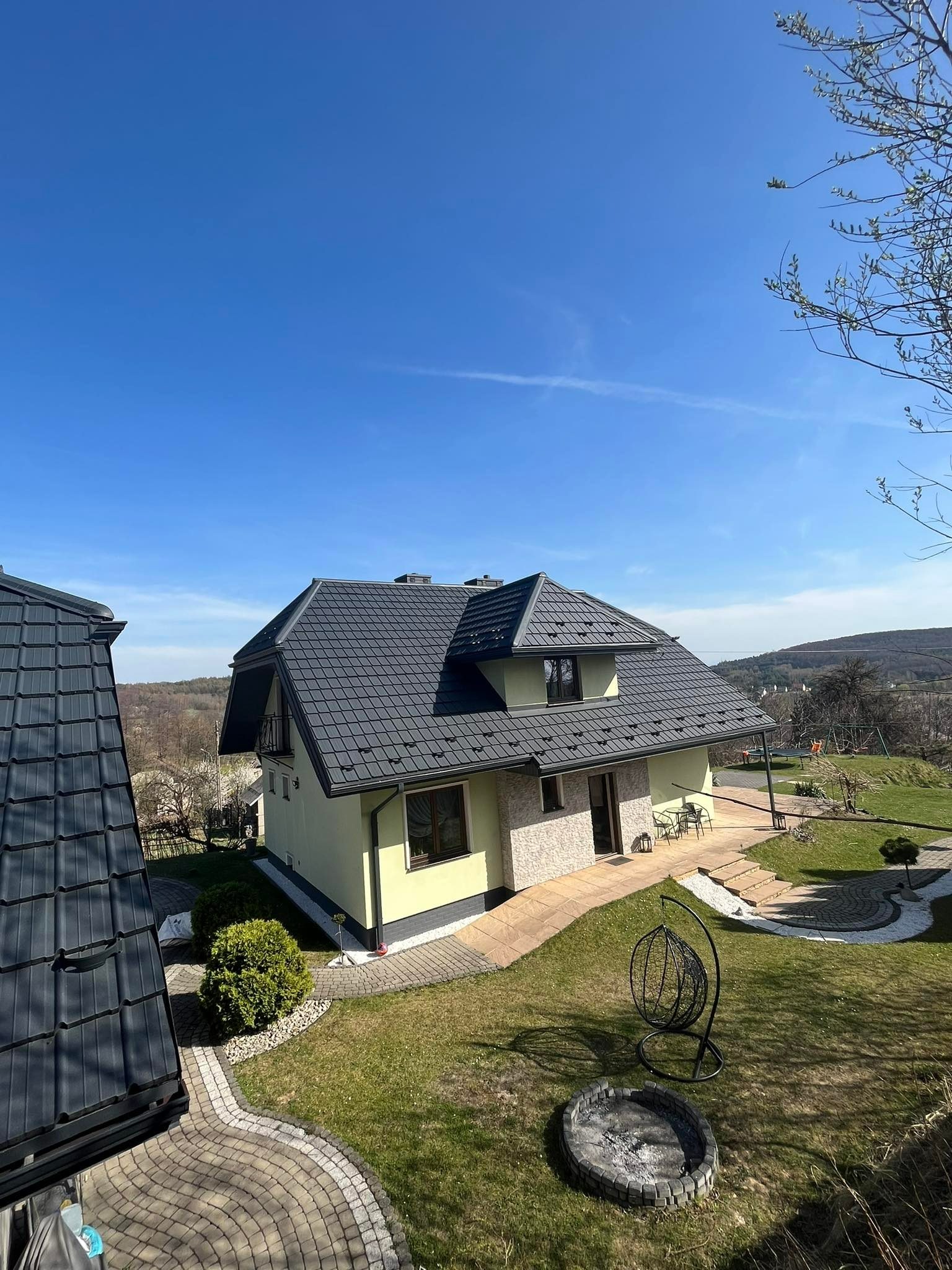 Dom z ciemnoszarą dachówką modułową, widok z góry, ogród z paleniskiem i wiszącym fotelem.