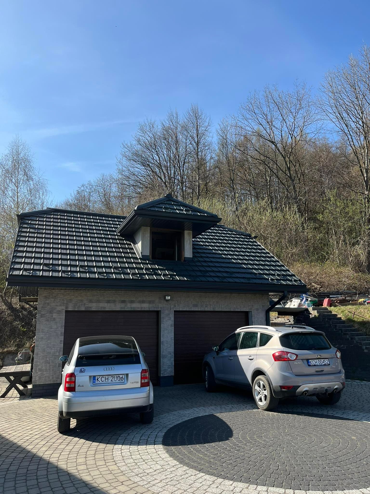 Dom z dwustanowiskowym garażem i ciemną dachówką ceramiczną, na podjeździe dwa zaparkowane samochody osobowe, w tle drzewa.