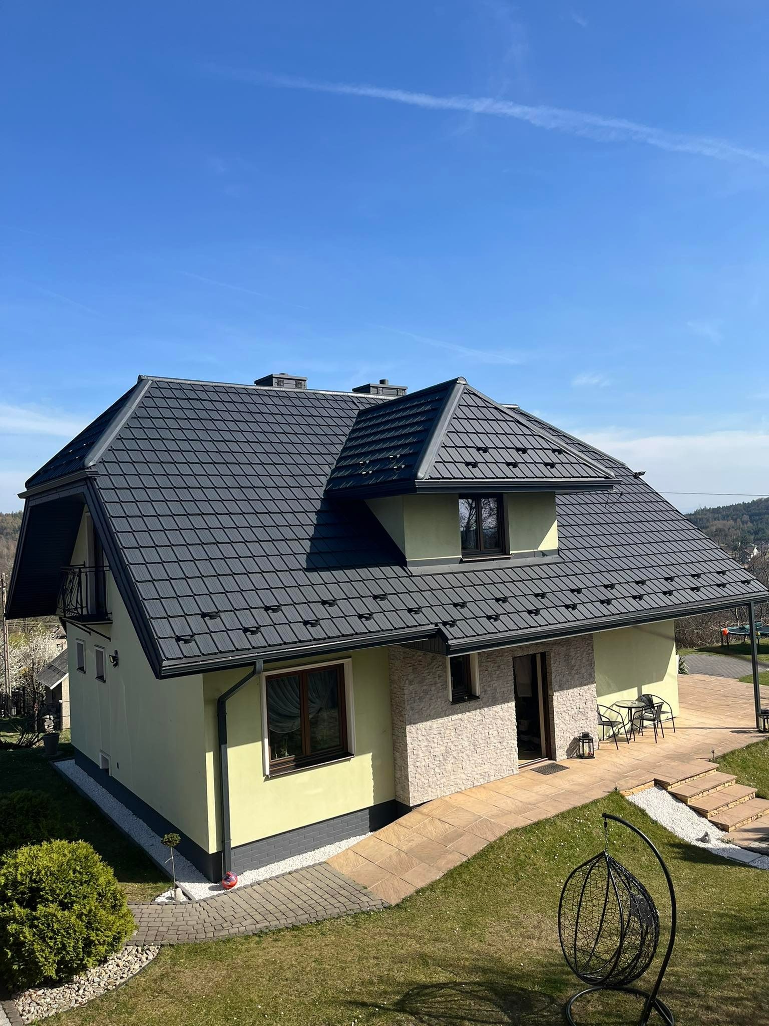 Nowoczesny dom jednorodzinny z grafitową blachodachówką, jasną elewacją i kamiennym wykończeniem, widok z góry na tle błękitnego nieba.