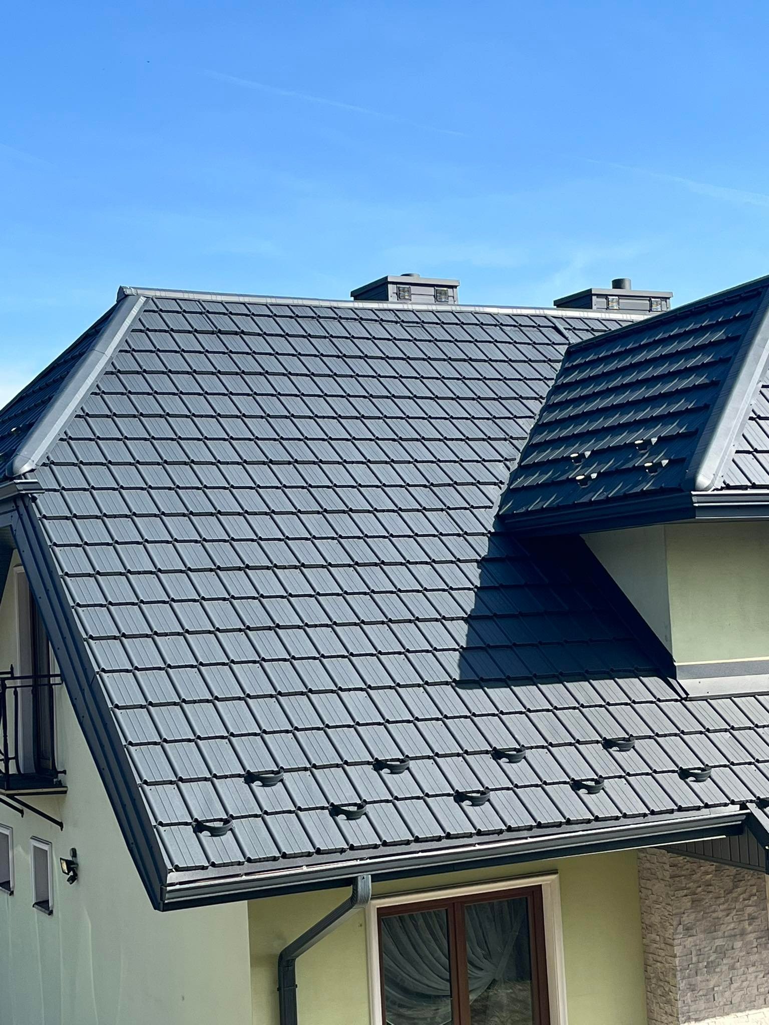 Szczegółowe ujęcie szarego dachu z dachówką karpiówką na jasnym domu z widocznymi kominami i rynnami w słoneczny dzień.