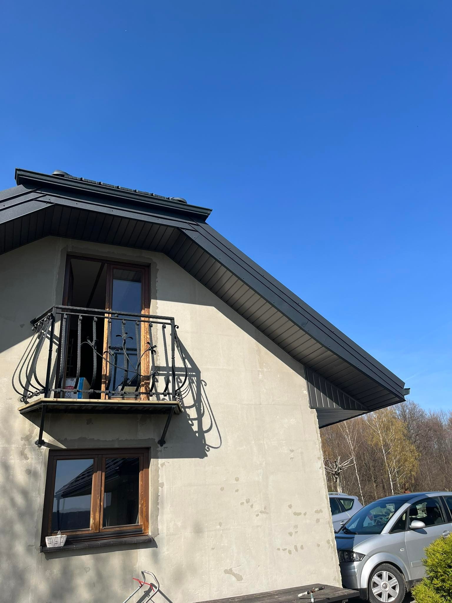 Dom w trakcie budowy lub remontu, widoczny balkon z ozdobną balustradą, okna z drewnianą ramą oraz fragment dachu z ciemną blachą. Elewacja surowa, nieotynkowana. W tle drzewa i zaparkowane samochody.