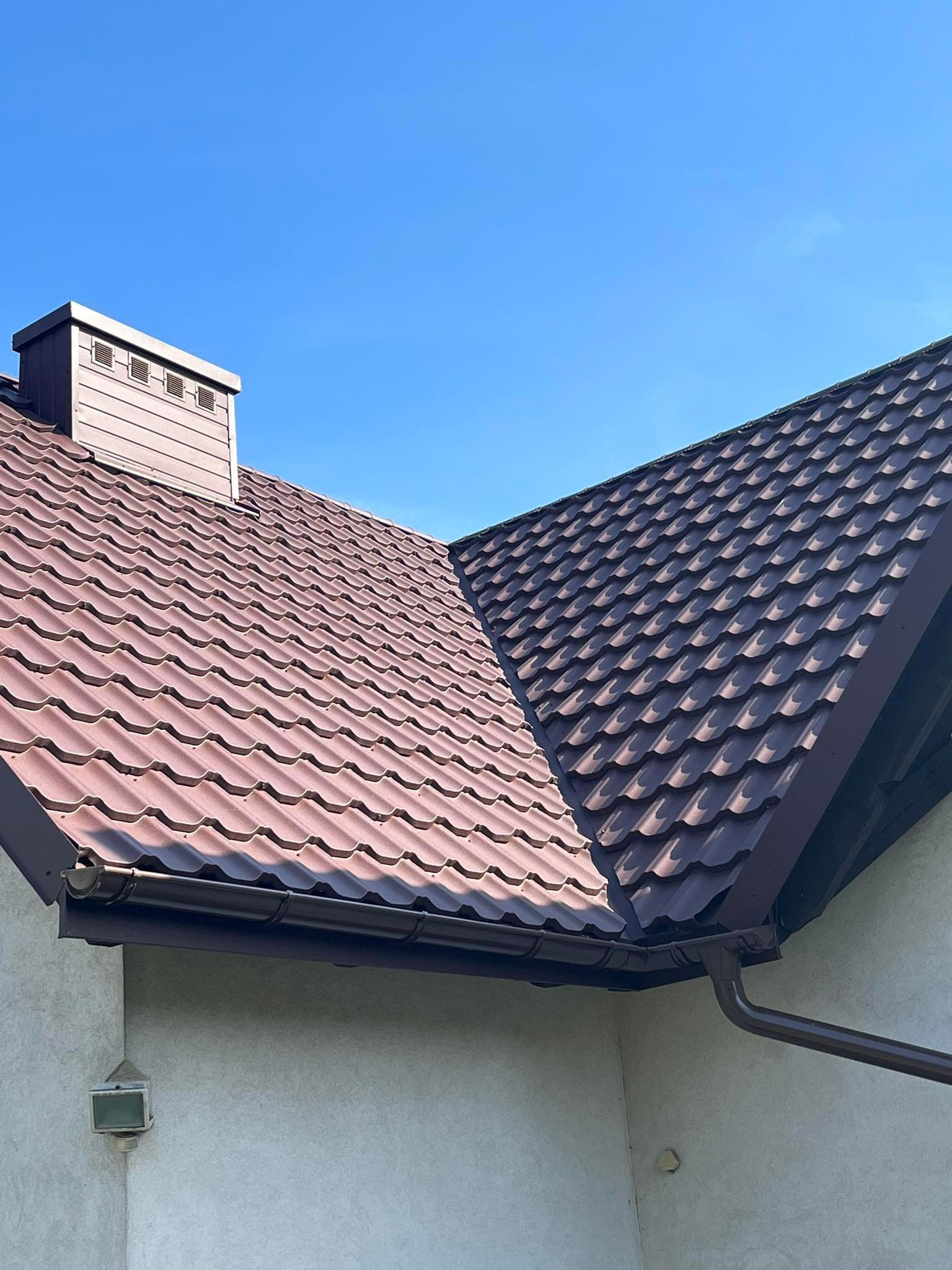 Brązowy dach z blachy trapezowej z kominem i rynnami, widok z dołu na tle błękitnego nieba.