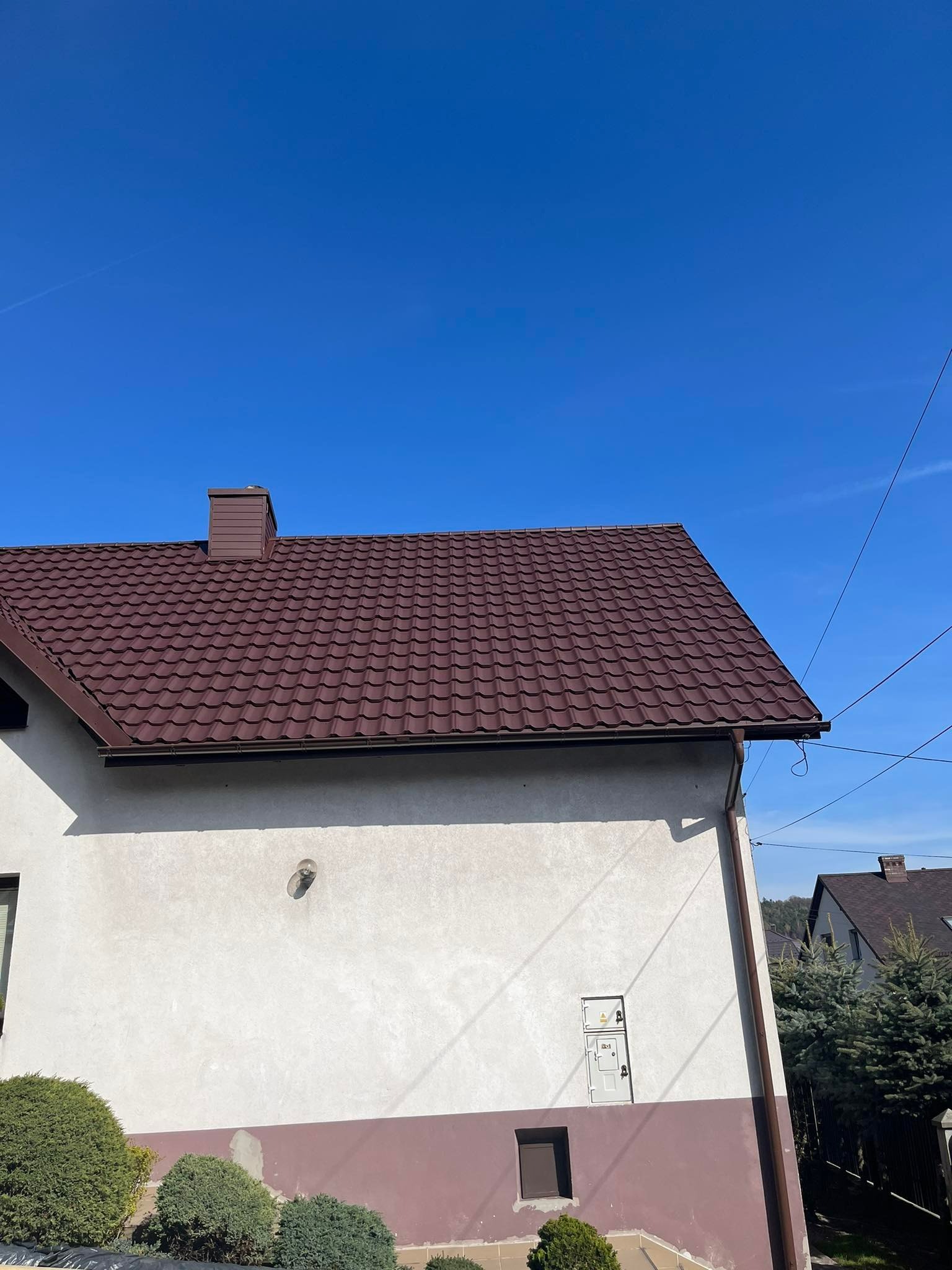 Dom jednorodzinny z nowym, brązowym pokryciem dachowym z blachy na tle błękitnego nieba, widoczne rynny i komin.