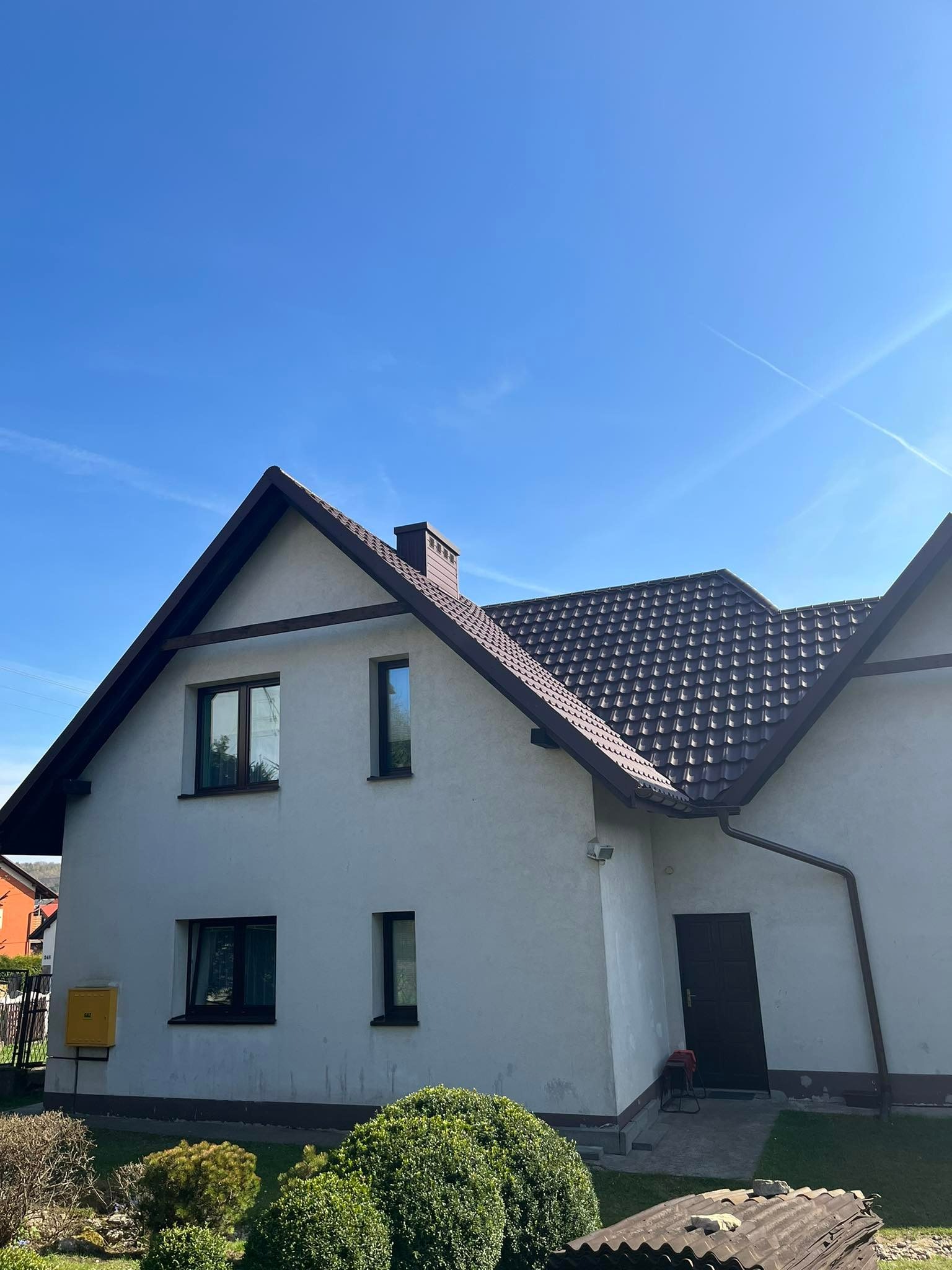 Dom jednorodzinny z ciemnobrązowym dachem z dachówki metalowej, brązowymi rynnami i białymi ścianami, widok z dołu na tle błękitnego nieba.