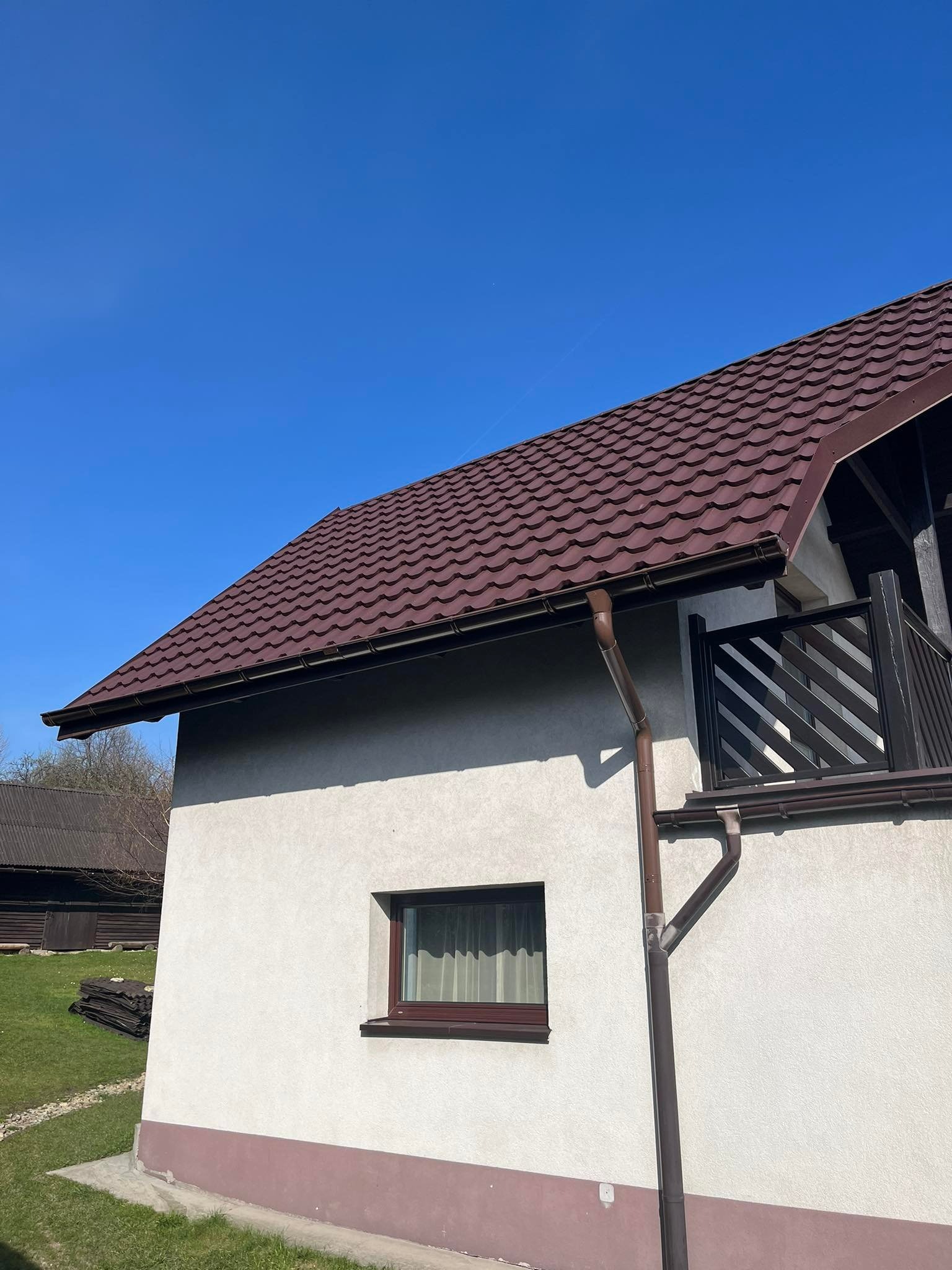 Dom jednorodzinny z brązowym dachem z blachodachówki, brązowymi rynnami i balkonem z drewnianą balustradą, widok z dołu na tle błękitnego nieba.