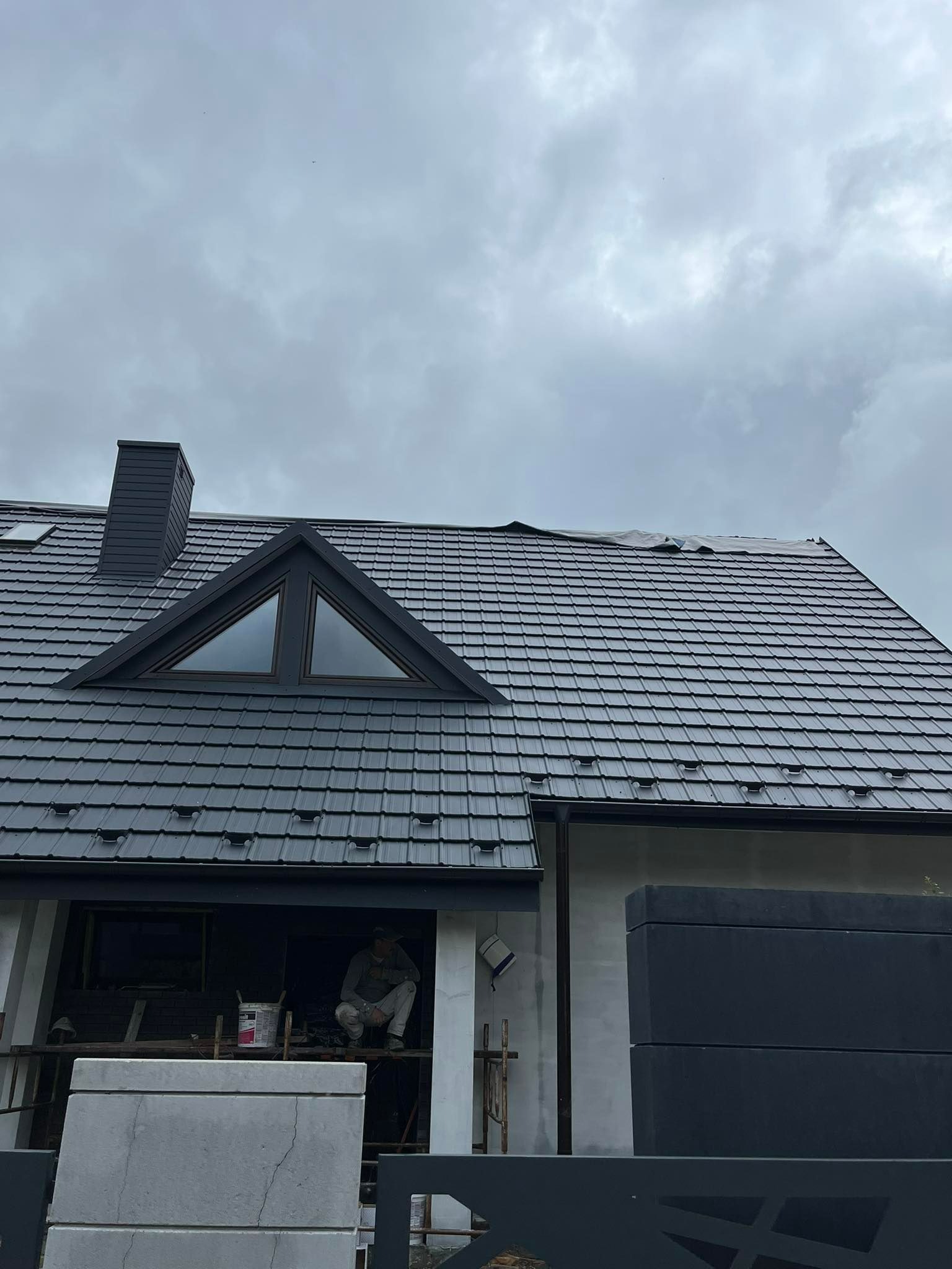 Montaż czarnej dachówki na nowym domu z oknem trójkątnym na poddaszu, dekarz pracuje na rusztowaniu.