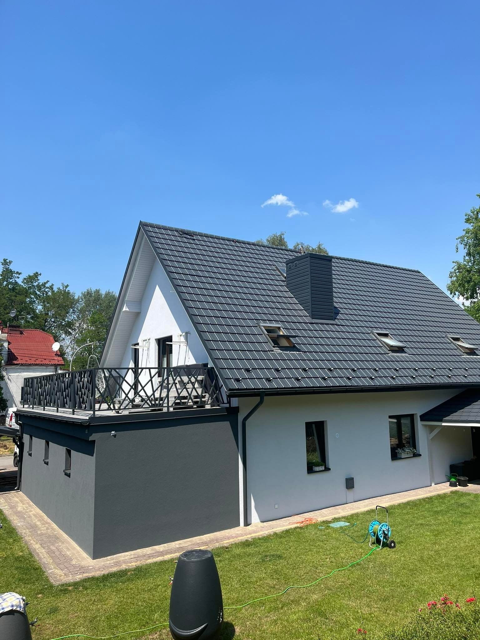 Nowoczesny dom jednorodzinny z szarą dachówką ceramiczną, balkonem z ażurową balustradą i zadbanym trawnikiem z wężem ogrodowym.