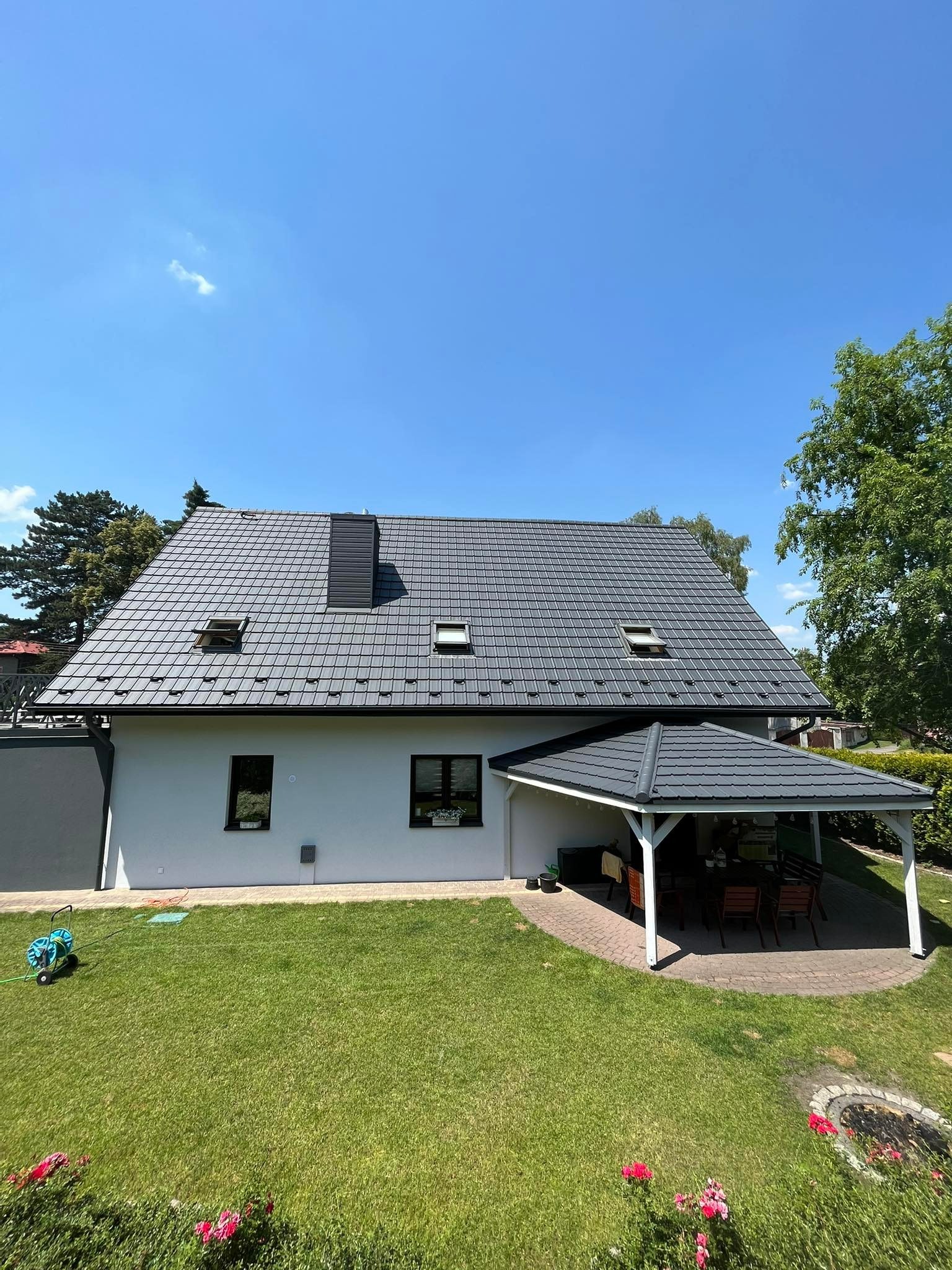 Dom jednorodzinny z szarym dachem z dachówki ceramicznej, widoczne okna dachowe, komin, biała elewacja, zielony trawnik i altana ogrodowa z szarym dachem.