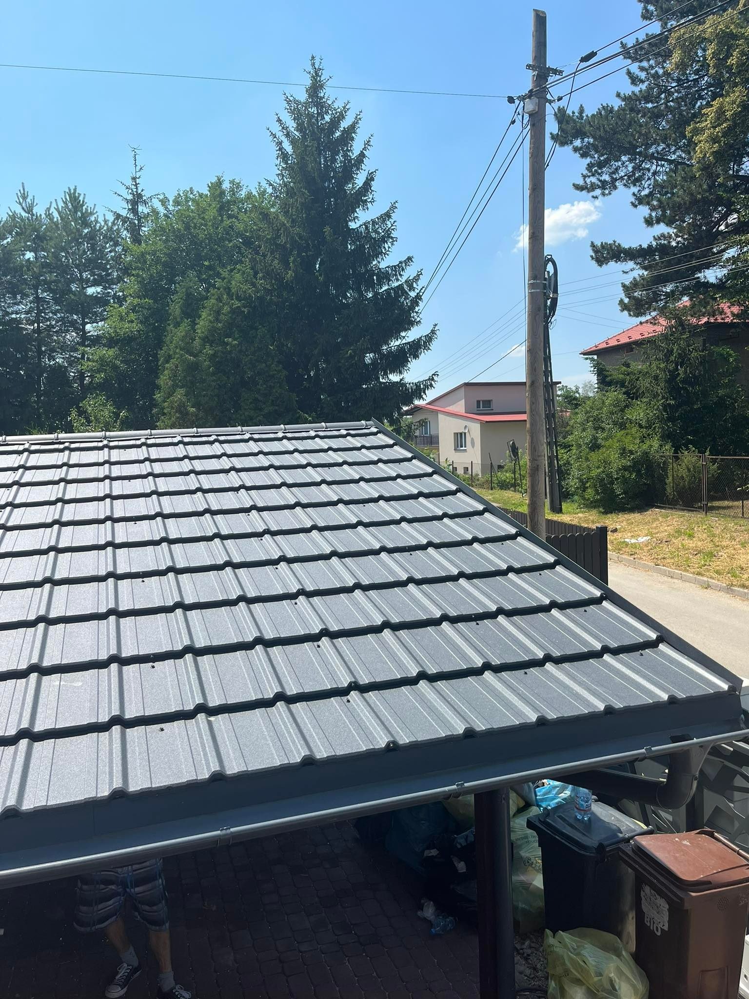 Nowy, szary dach z metalowej dachówki modułowej, widok z dołu, w tle drzewa, słup elektryczny i budynki.