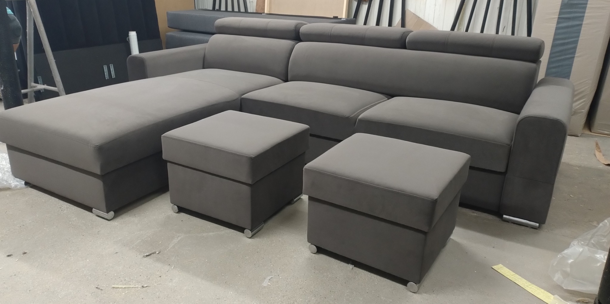 Szara, modułowa sofa narożna z dwoma pufami na metalowych nóżkach, ustawiona w magazynie przed wysyłką.
