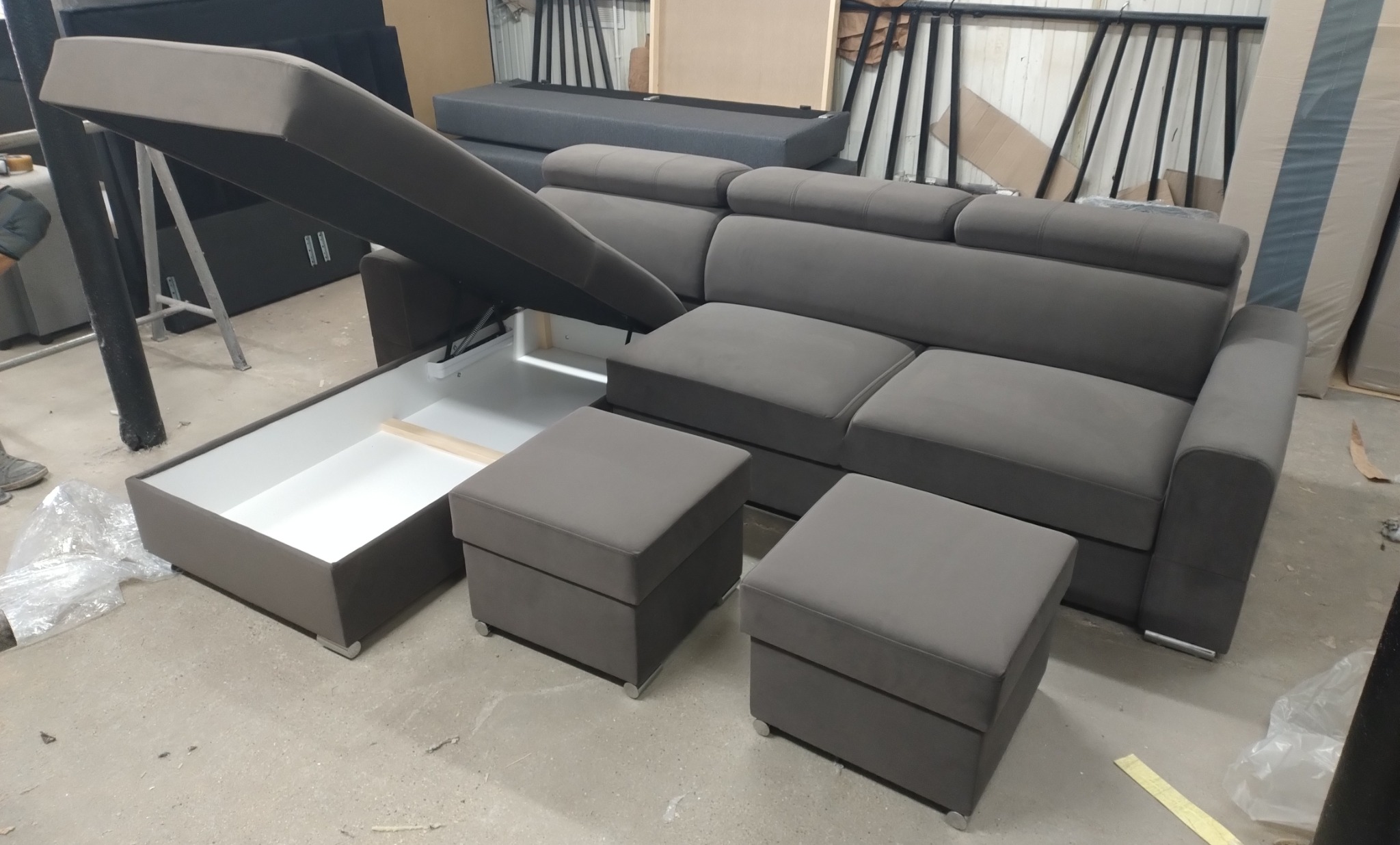 Szary narożnik z podnoszonym siedziskiem, ukazującym biały pojemnik na pościel; dwa pufy w kolorze narożnika, surowe wnętrze warsztatu w tle.