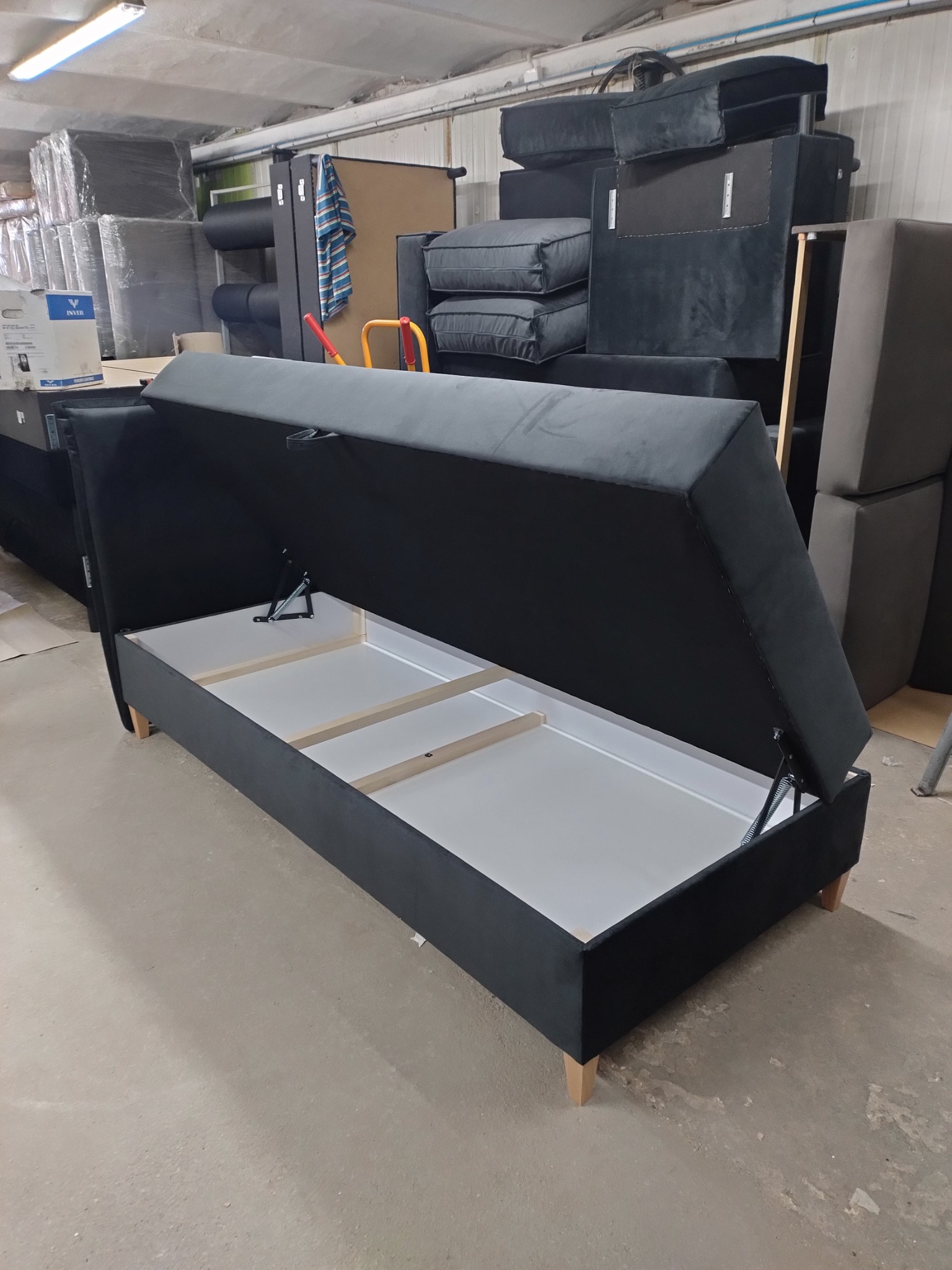 Czarne tapicerowane łóżko z podnoszonym pojemnikiem na pościel, widoczne mechanizmy podnoszące, w tle magazyn mebli w Kostowie.