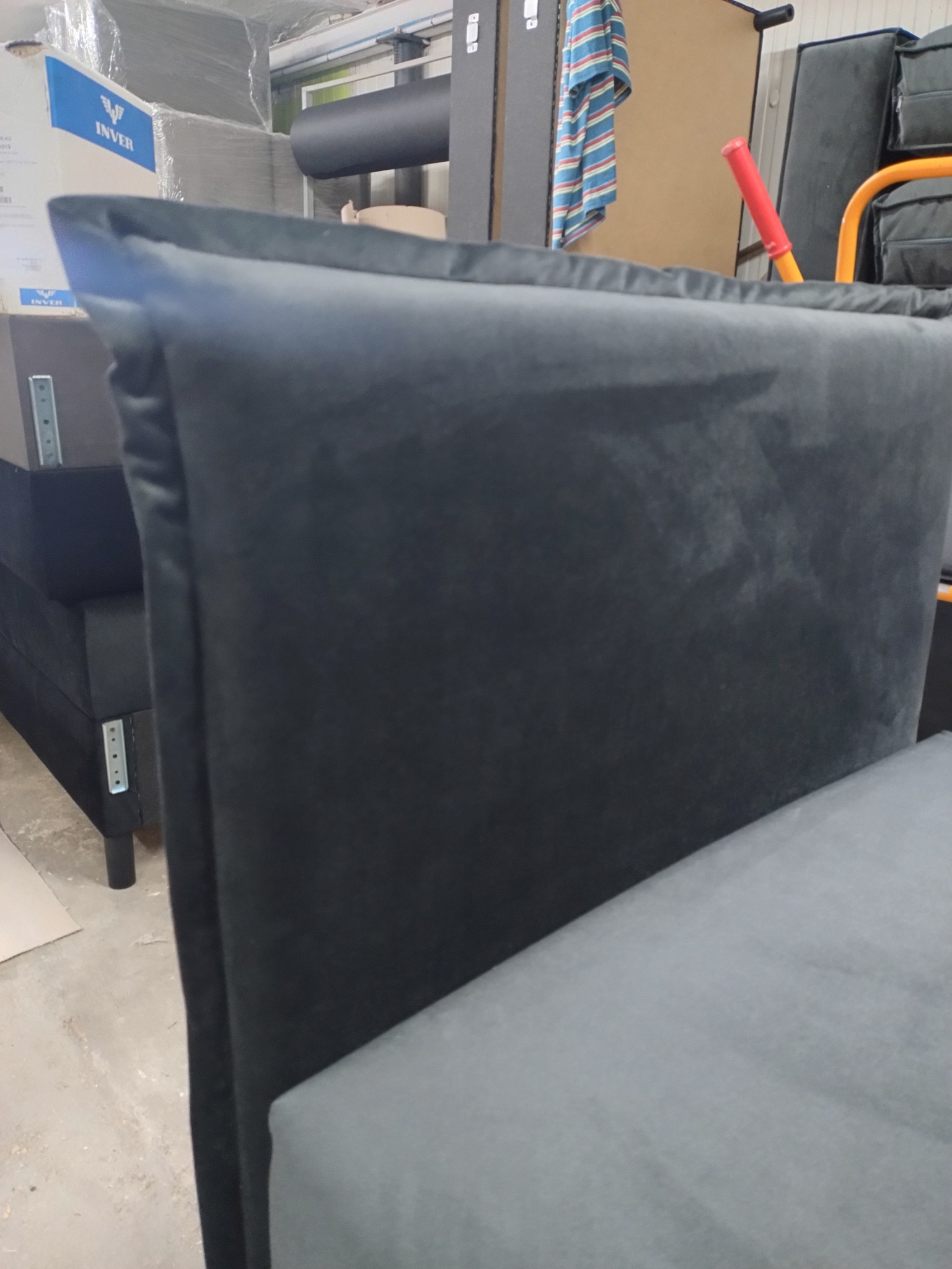 Fragment ciemnoszarego, pluszowego zagłówka łóżka z widocznym fragmentem siedziska, na tle magazynu z zapakowanymi towarami i elementami wyposażenia.