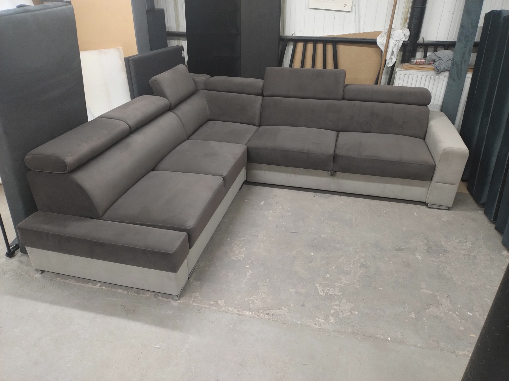 Szary narożnik z regulowanymi zagłówkami, widoczny w magazynie lub warsztacie, na betonowej podłodze.