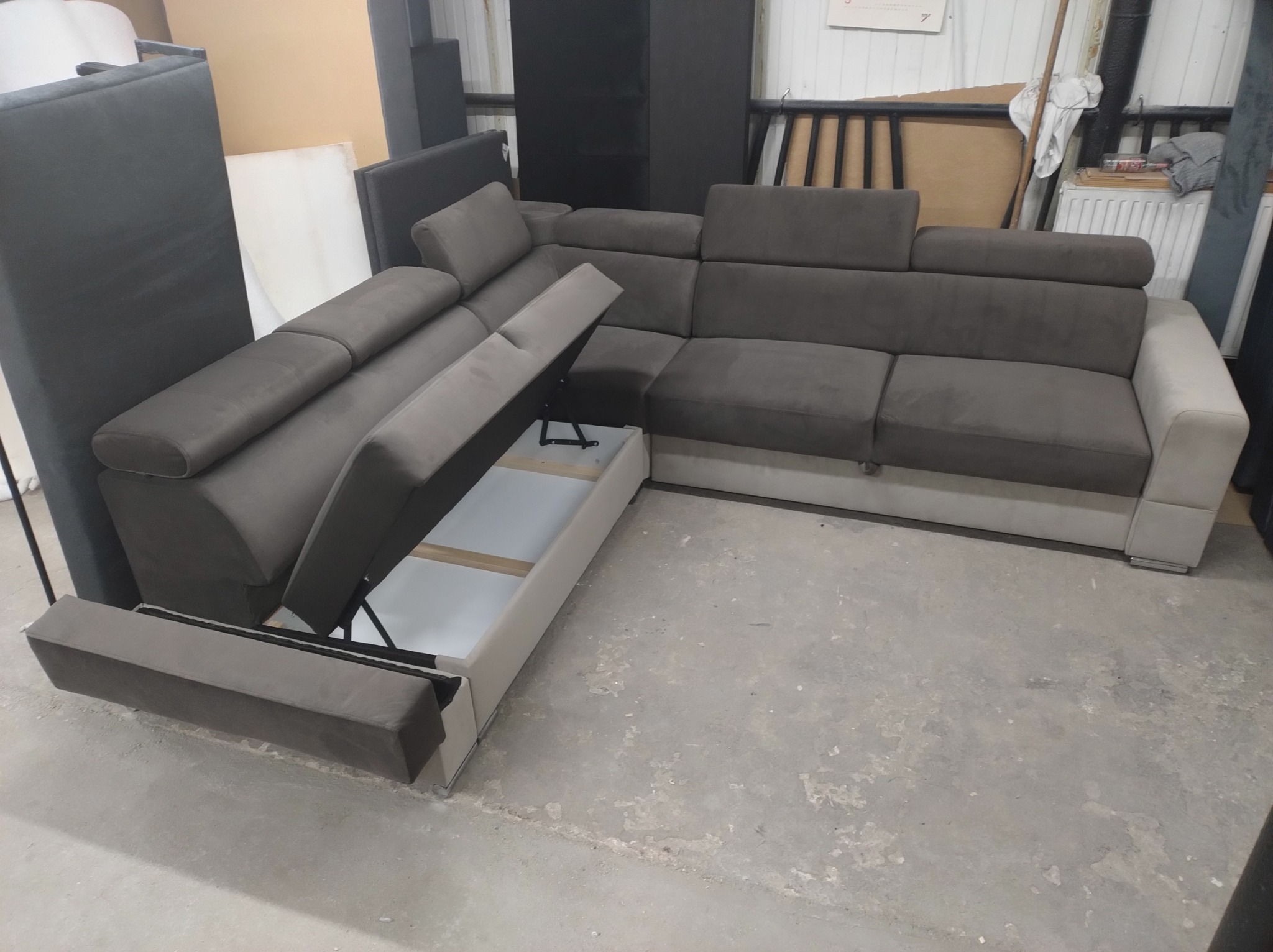 Szary narożnik z podnoszonym siedziskiem, ukazującym biały pojemnik na pościel, na tle surowego, betonowego podłoża i elementów wyposażenia magazynowego.