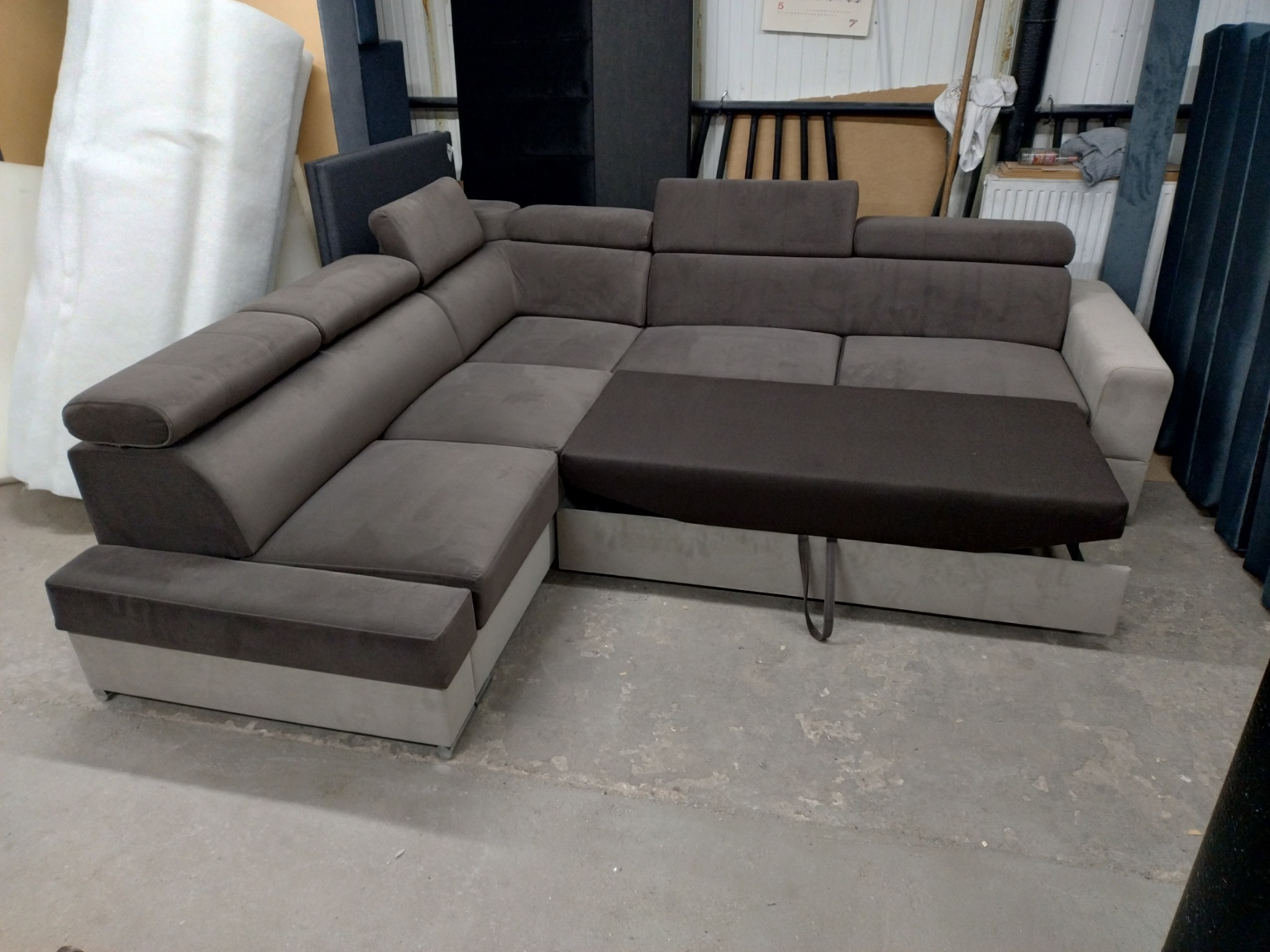 Rozłożony narożnik z funkcją spania w odcieniach szarości i brązu, widoczny mechanizm wysuwania, w tle materiały i elementy wyposażenia warsztatu.