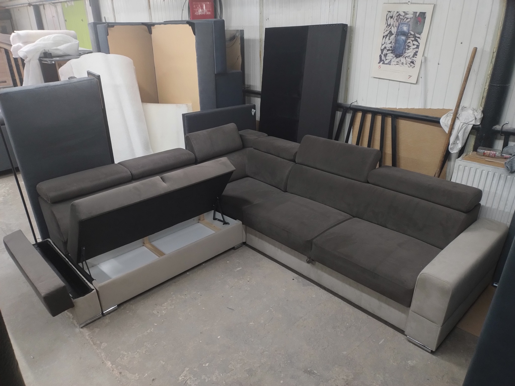 Szary narożnik z otwartym pojemnikiem na pościel i podnoszonym bokiem, widoczne elementy konstrukcyjne, tło magazynowe z materiałami tapicerskimi.