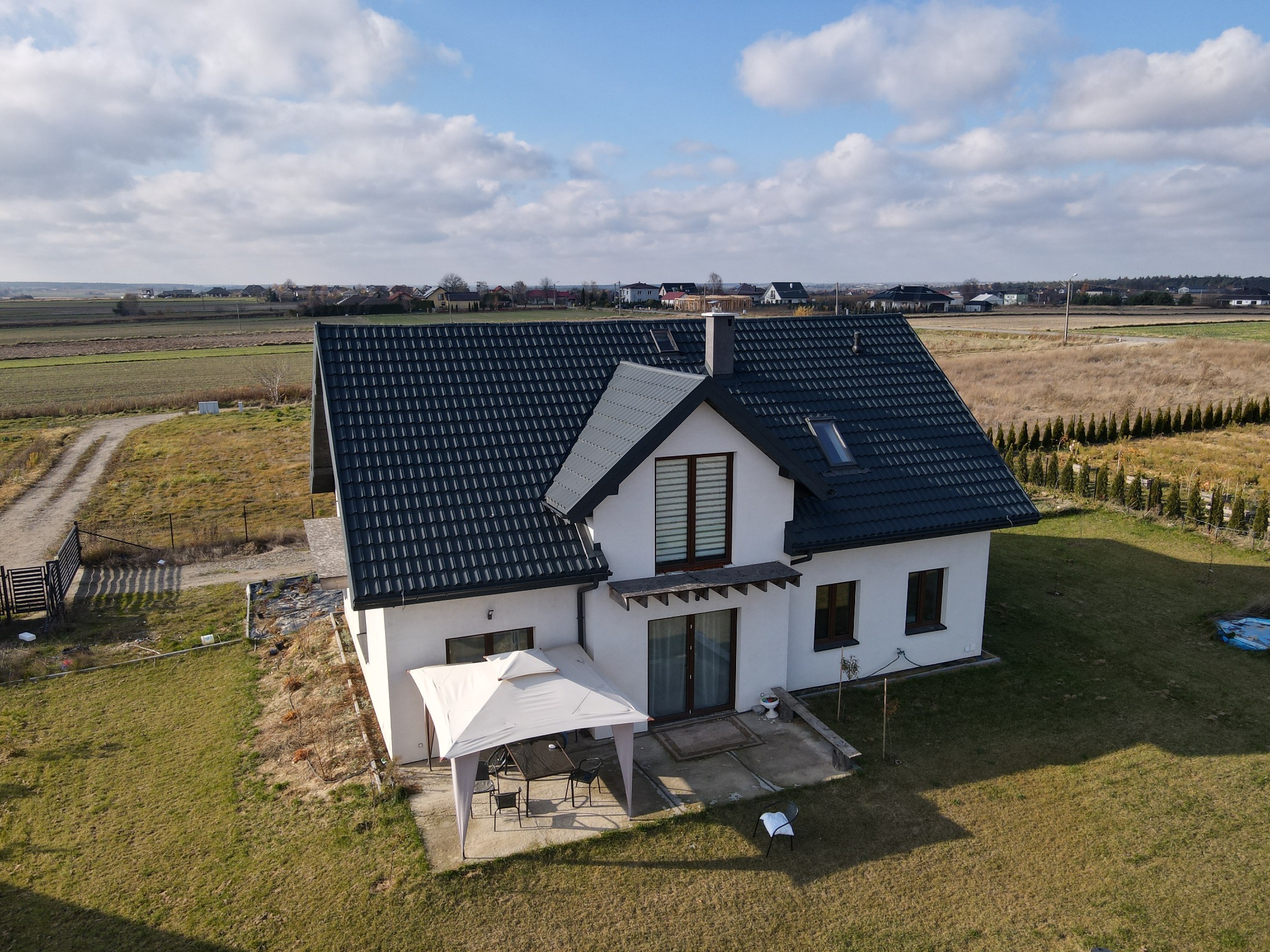 Nowoczesny, biały dom jednorodzinny z ciemną dachówką, widziany z lotu ptaka, z altaną ogrodową na tarasie i rozległym terenem wokół.