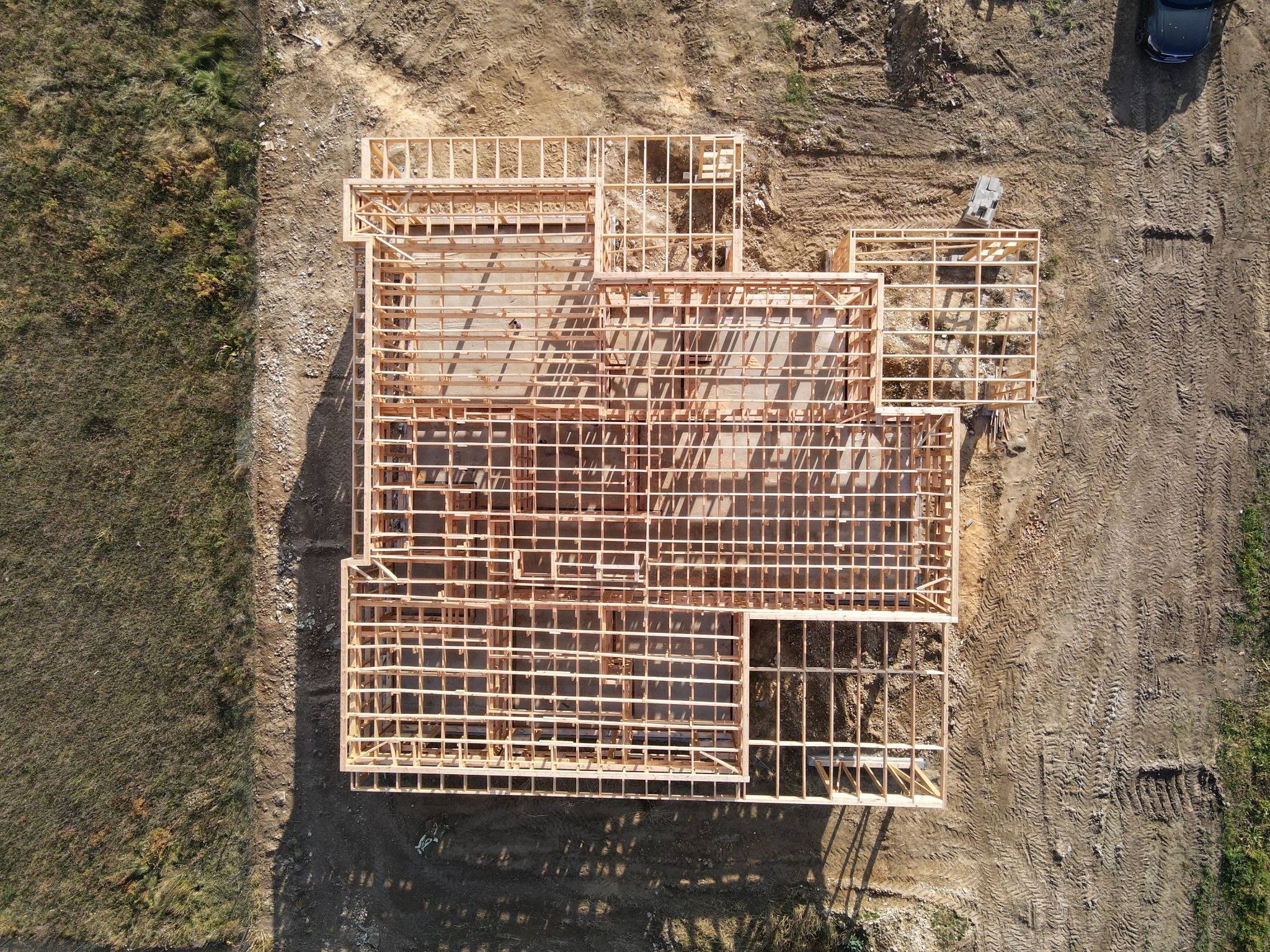 Widok z drona na drewnianą konstrukcję domu w trakcie budowy, z widocznymi fundamentami i zarysem ścian na nierównym terenie budowy.