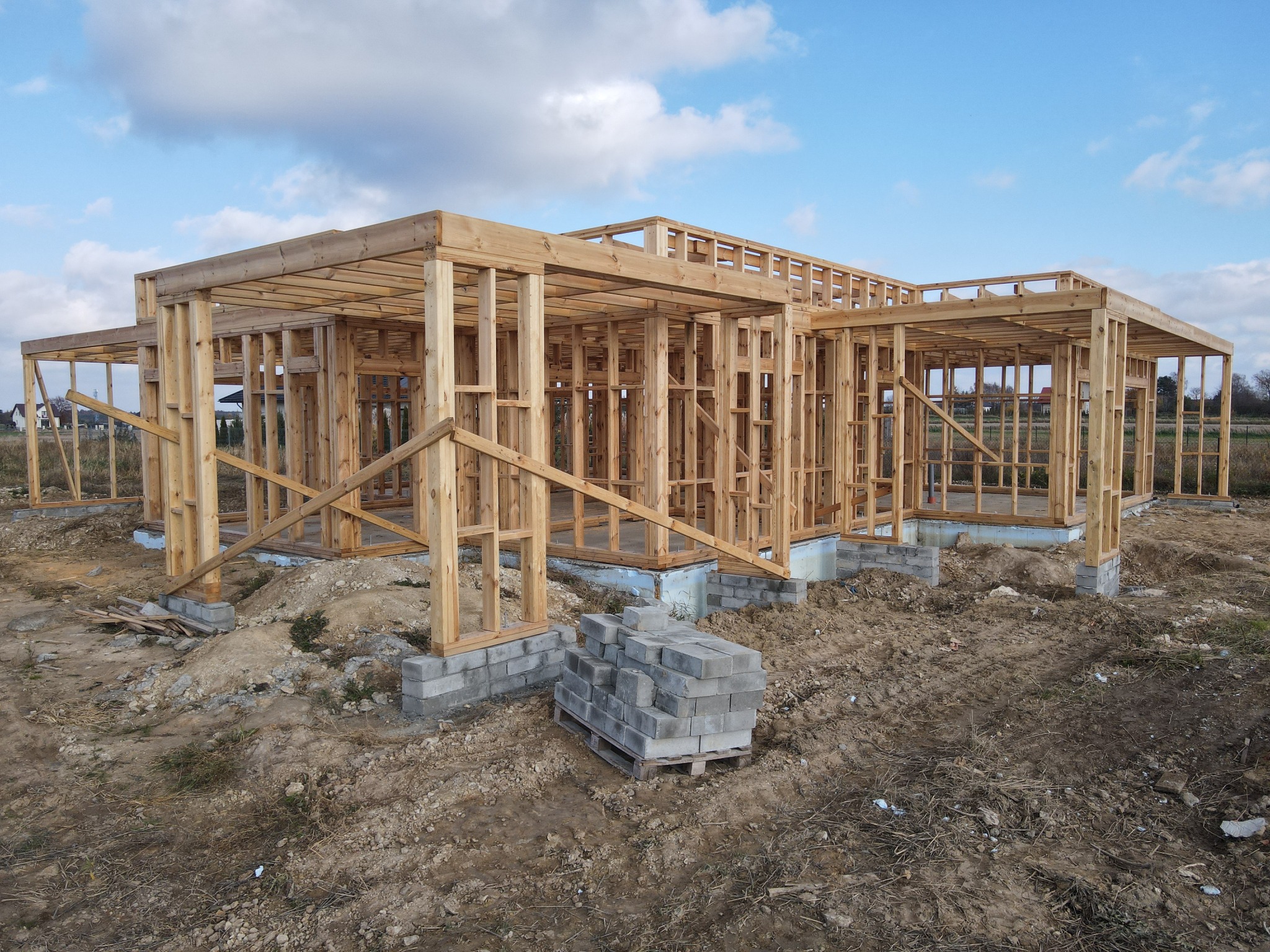 Surowy szkielet drewnianego domu w trakcie budowy, widoczne fundamenty i stos szarych bloczków betonowych na palecie, niebo z chmurami w tle.